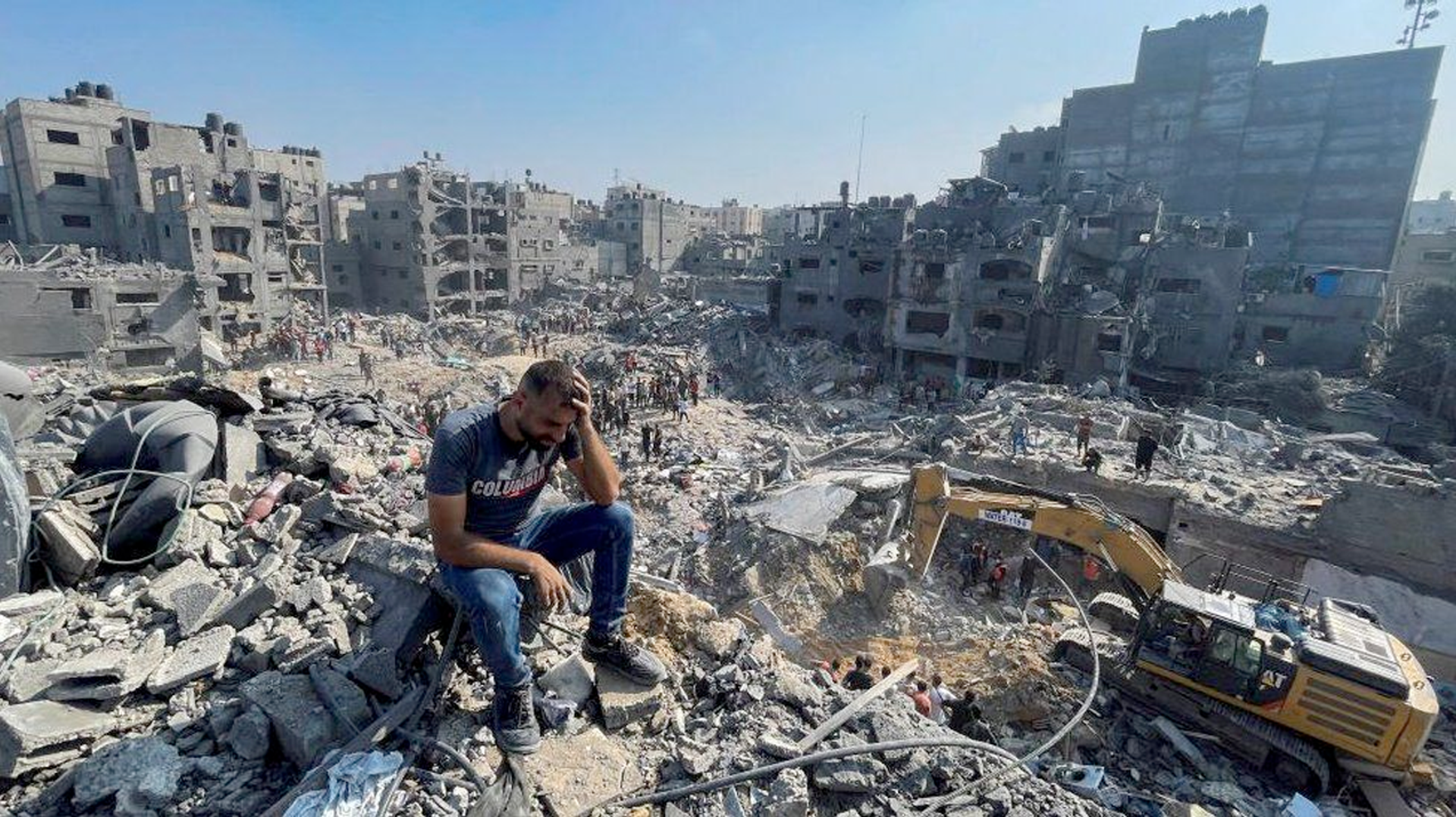 A man sits on a pile of rubble looking depressed as machinary cleans up the destroyed town Nov 3rd, 2023