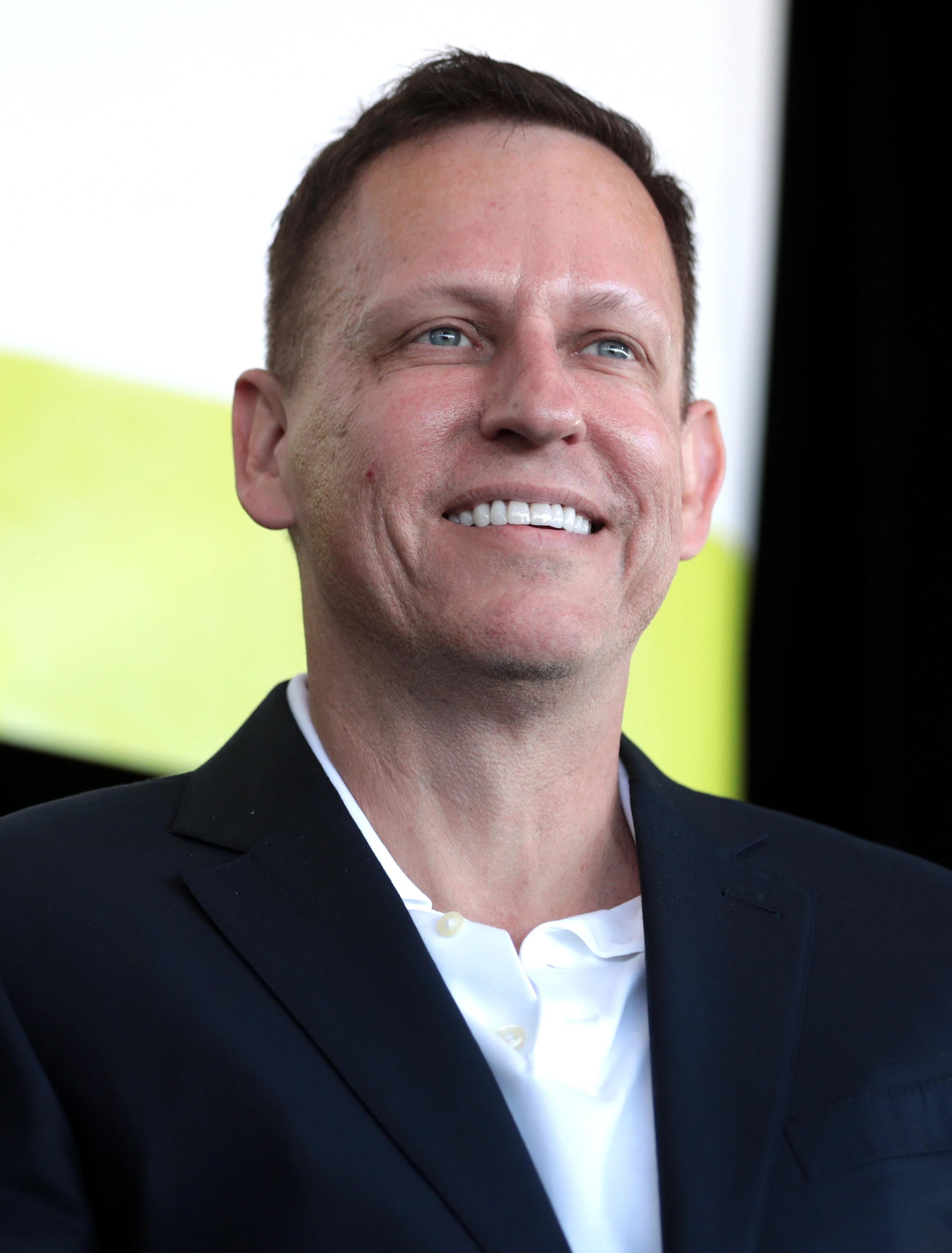 Peter Thiel, a middle-aged man with short dark hair, wearing a dark suit and tie, speaking at a podium at a political event