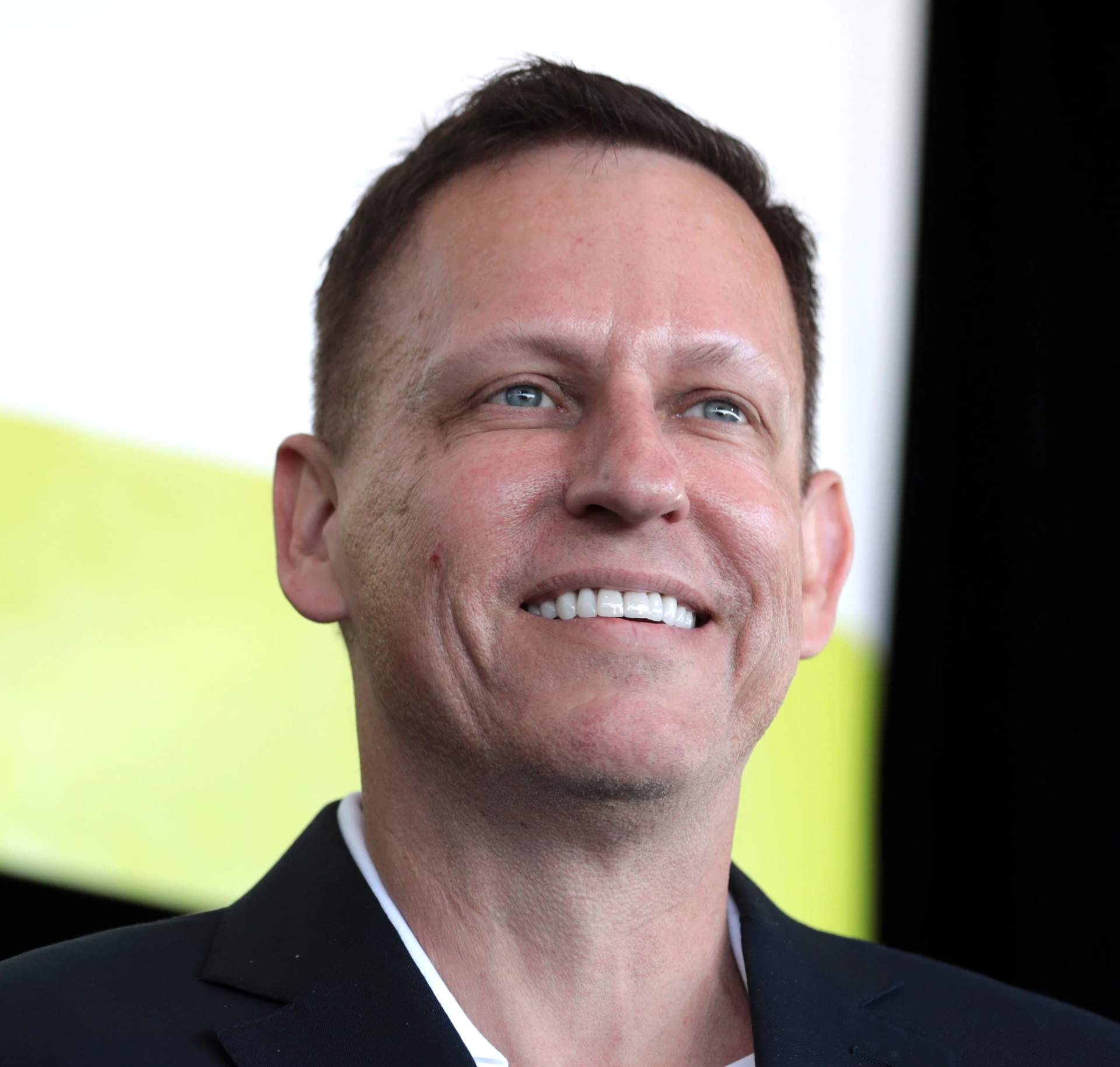 Peter Thiel, a middle-aged man with short dark hair, wearing a dark suit and tie, speaking at a podium at a political event
