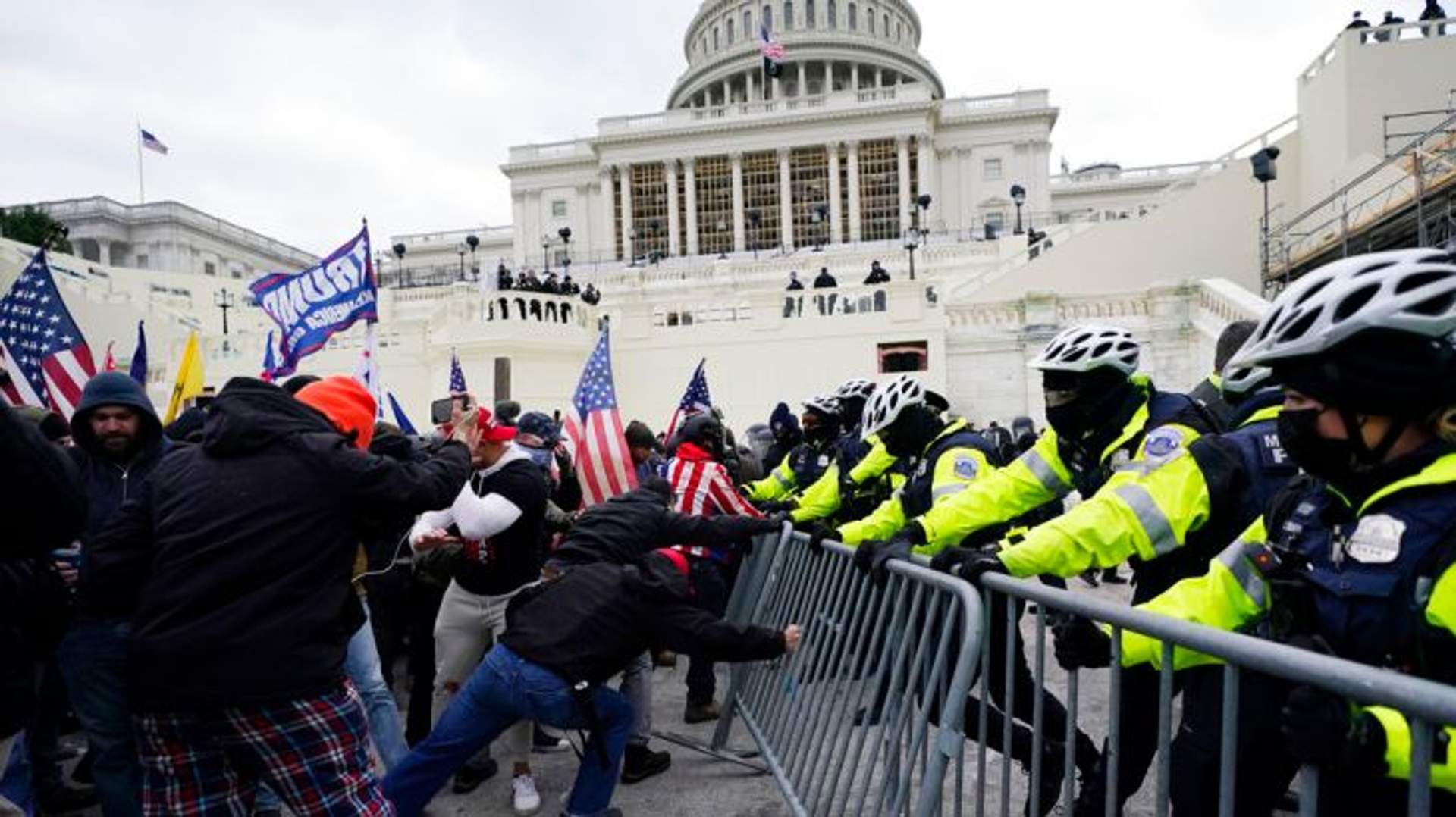 A chaotic scene at the U.S. Capitol building on January 6, 2021, showing a large crowd of pro-Trump supporters breaching security barriers and climbing the Capitol steps under overcast skies.