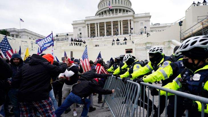 A chaotic scene at the U.S. Capitol building on January 6, 2021, showing a large crowd of pro-Trump supporters breaching security barriers and climbing the Capitol steps under overcast skies.