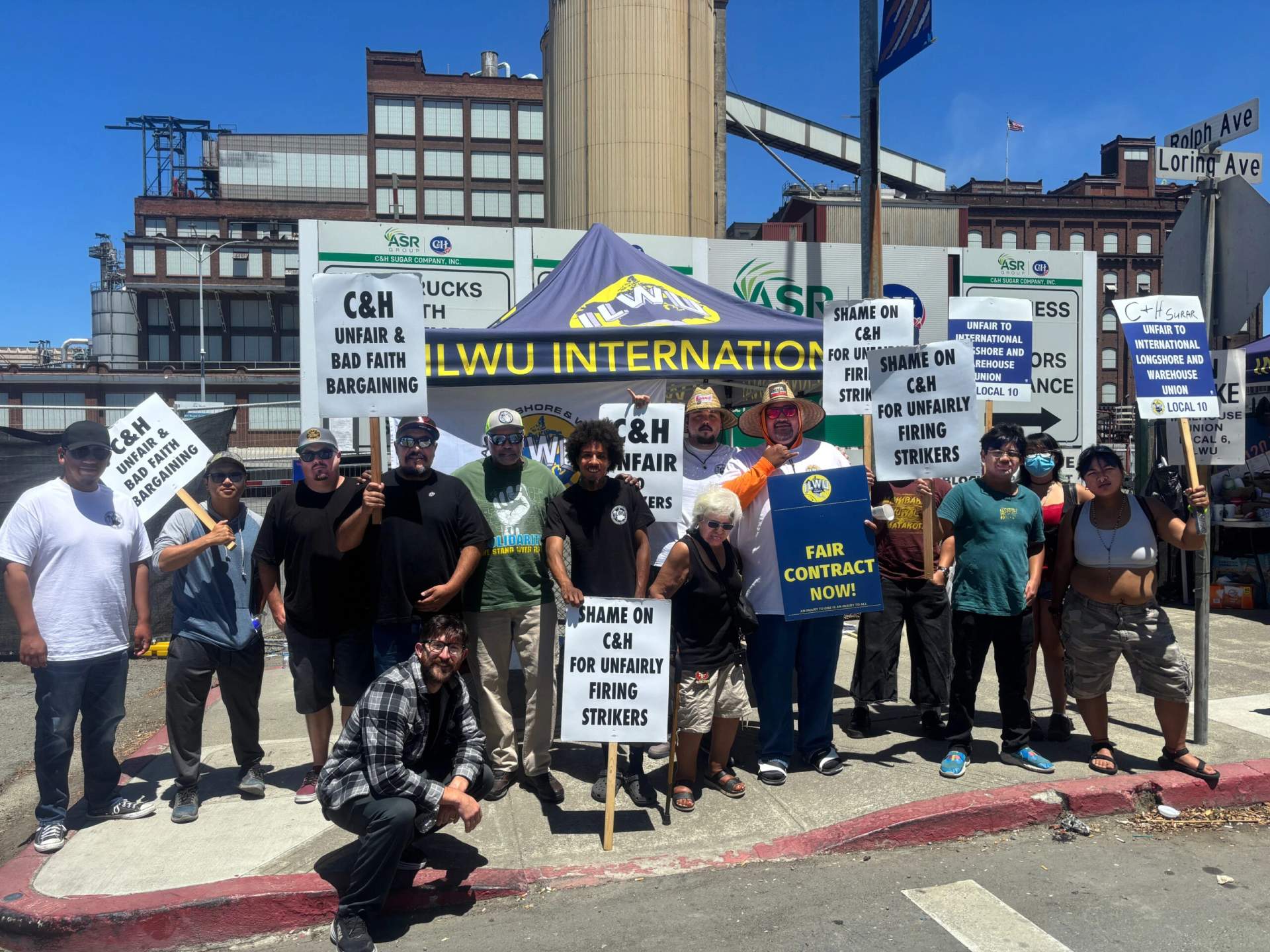 A group of ILWU members and supporters stand on a street holding signs and union banners outside an industrial refinery with a water tower in the background in Crockett California