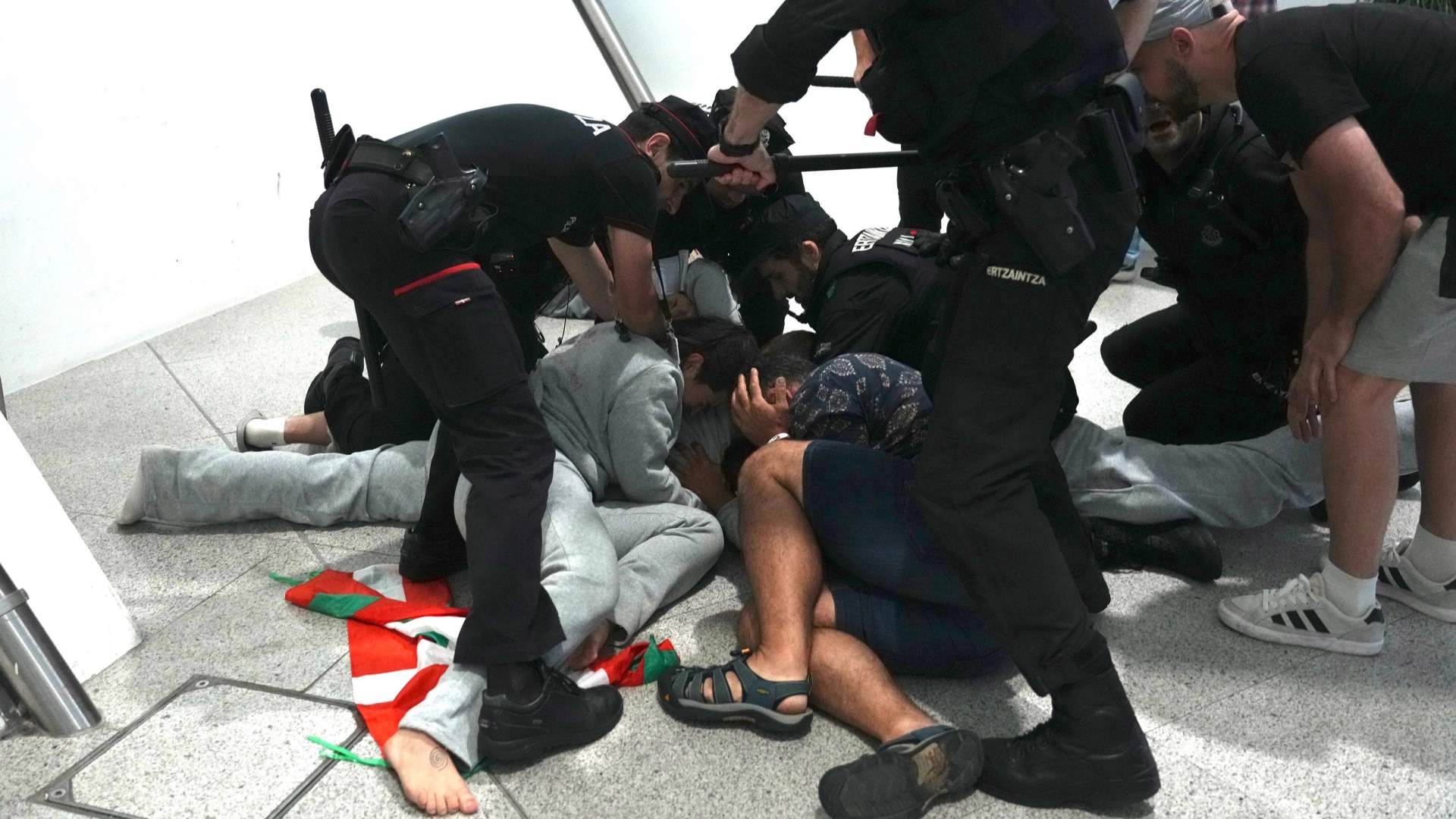 Basque Ertzaintza police officers swinging batons at pro-Palestinian activists on the floor of Bilbao airport arrivals hall. Several people are on the ground. Others are scrambling away.