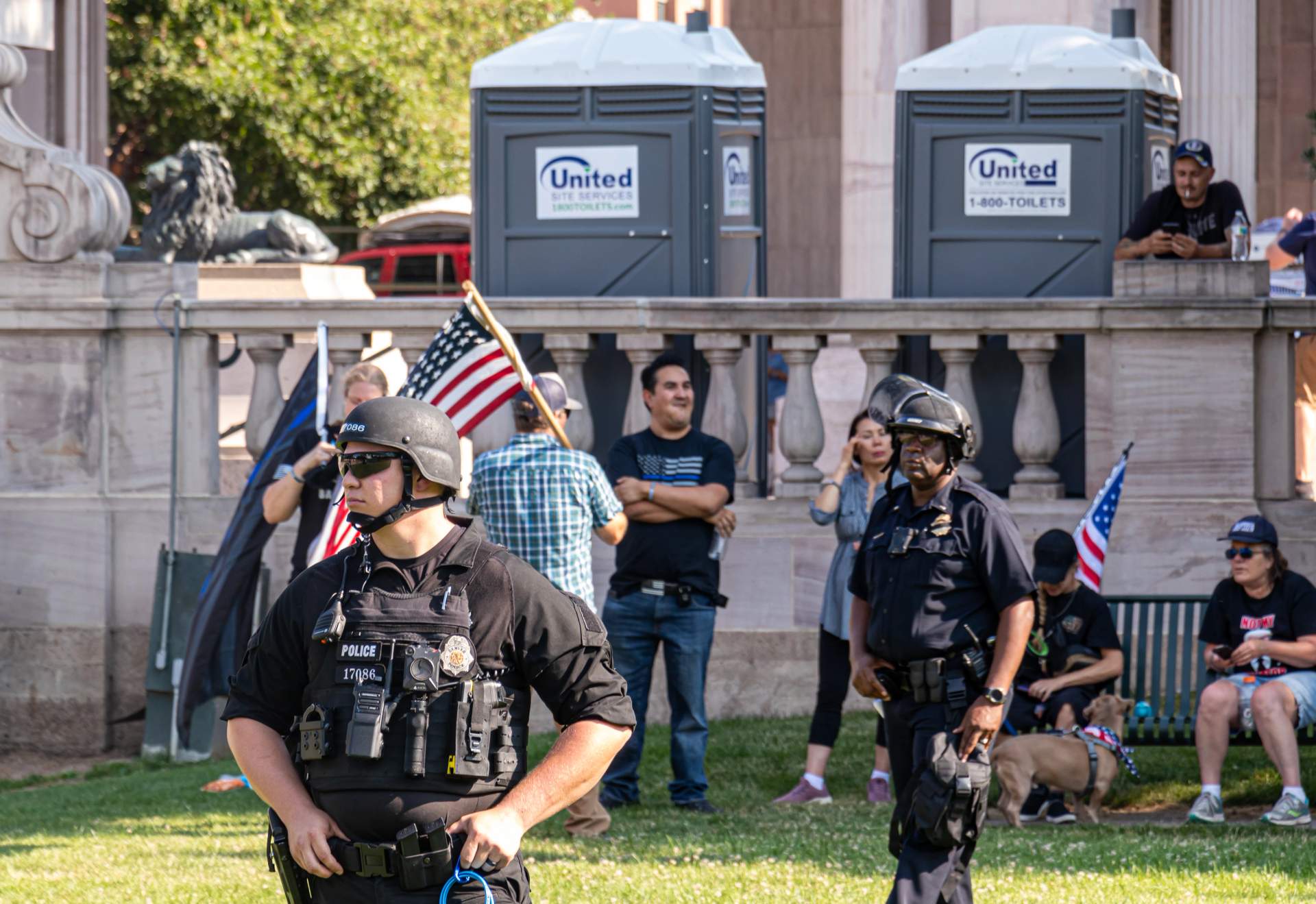 Through the chain-link: the police line faces outward, their backs to the people celebrating them. The architecture of control — fence on the outside, armor on the inside, the people they've hurt on the other side.