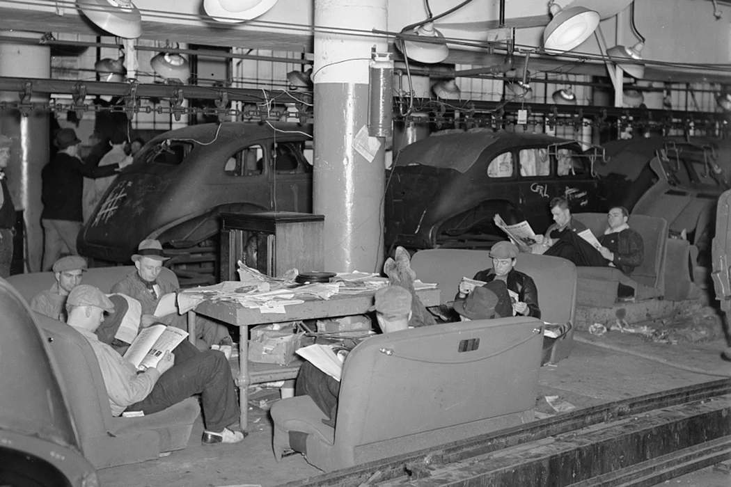 Men sitting on the factory floor inside an automobile manufacturing plant during the 1936-1937 Flint sit-down strike, with machinery and windows visible in the background
