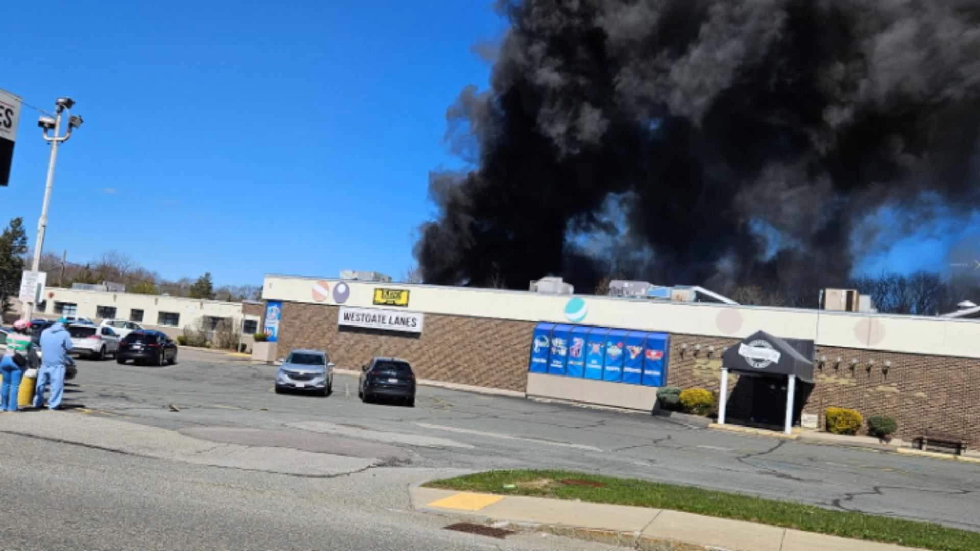 View from across a parking lot showing thick black smoke billowing from behind a bowling alley.