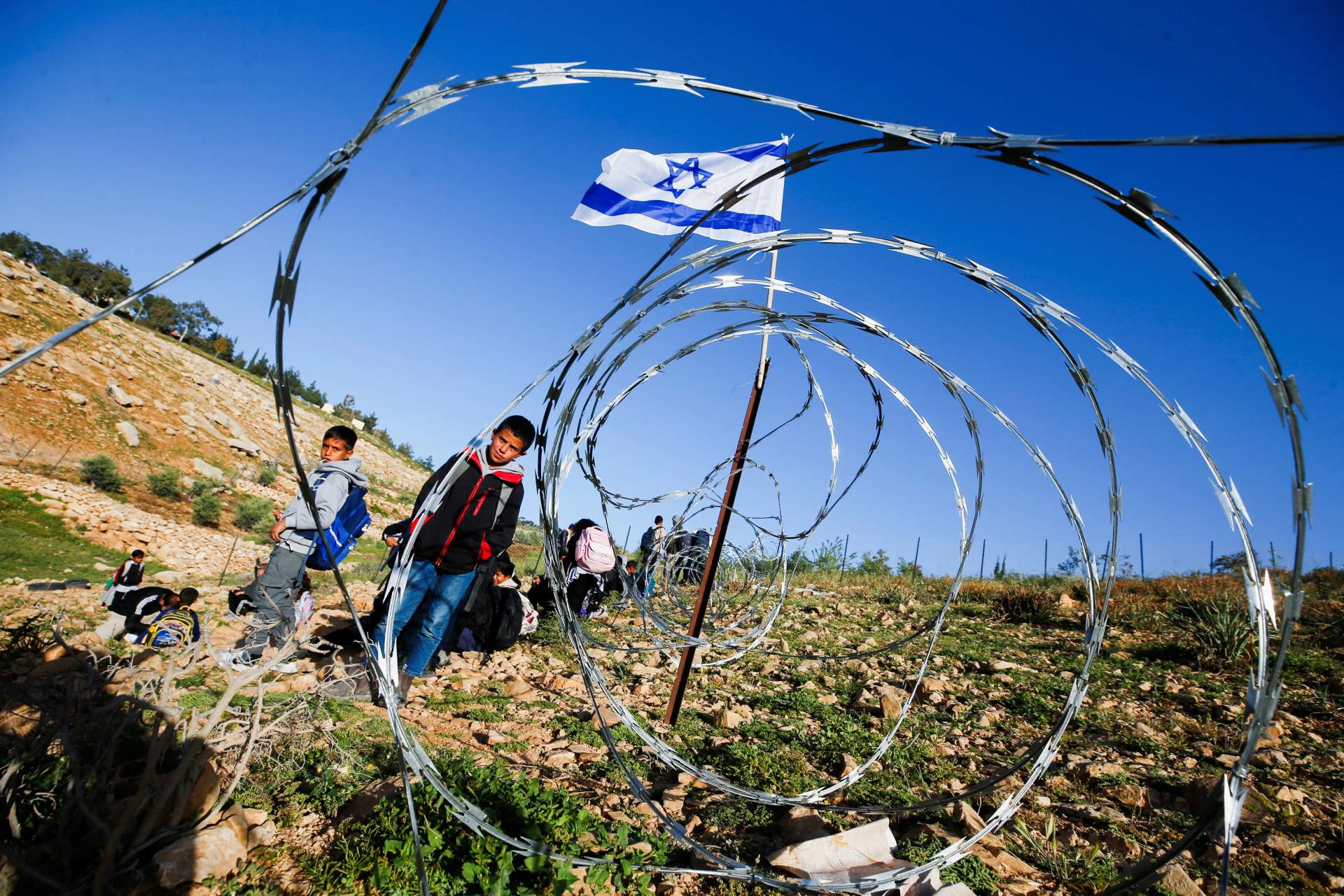 A group of Palestinian children and adults standing near a barbed-wire fence across a dirt road in an arid landscape in the occupied West Bank