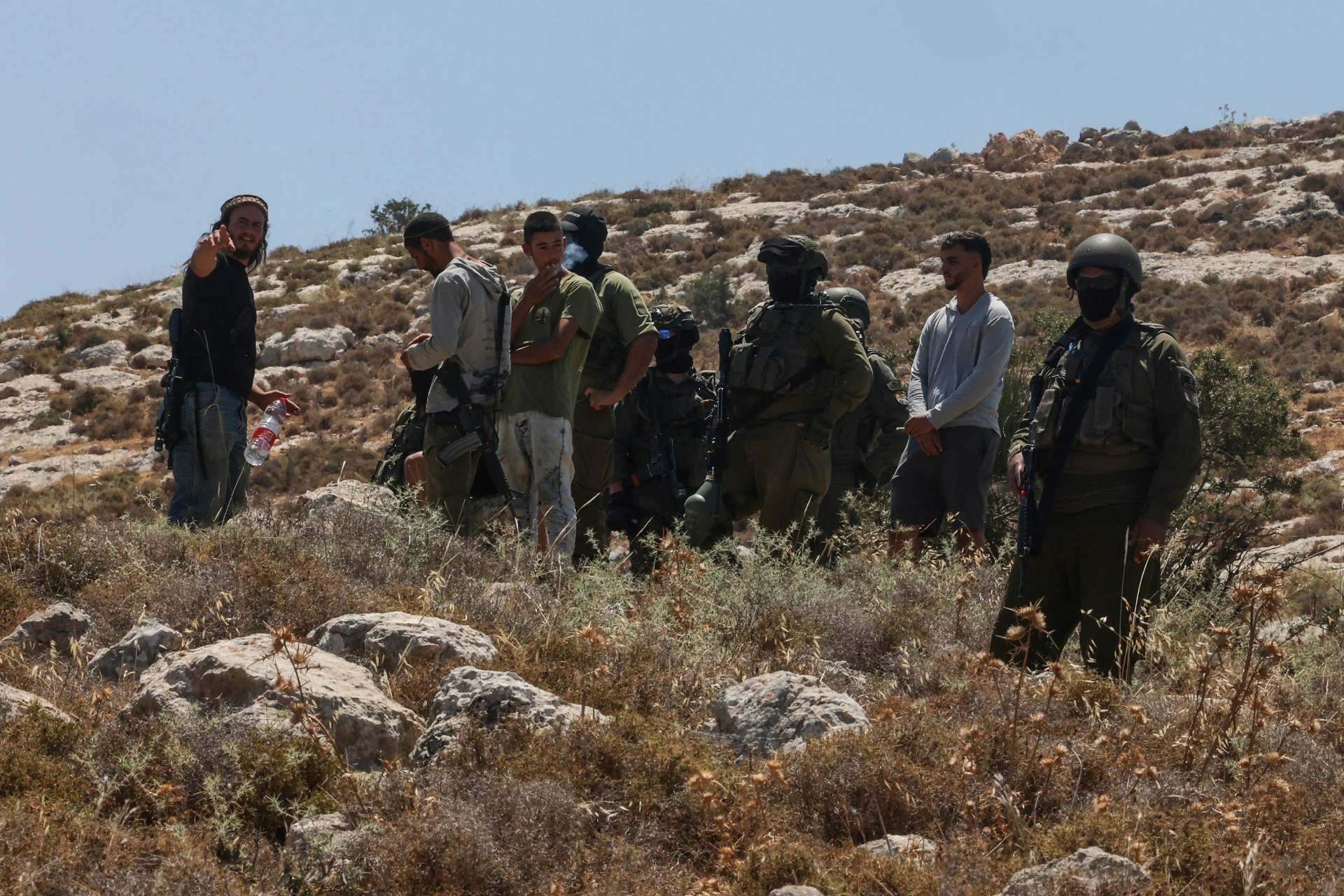 Armed men in military and civilian clothing standing near a bulldozer in a dusty landscape in the occupied West Bank