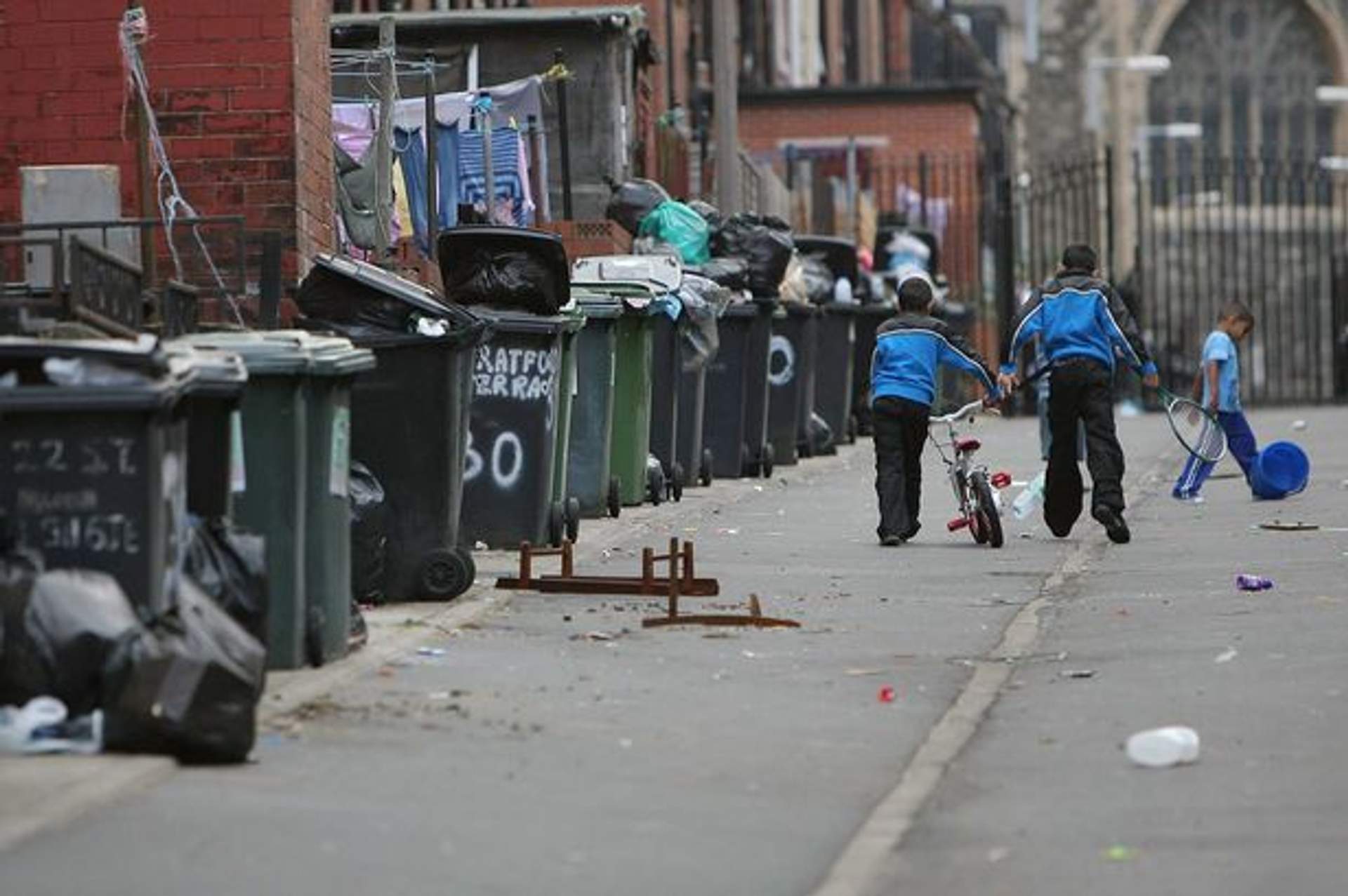 Overflowing rubbish bins line a residential street in the UK as two children walk past with a bicycle.