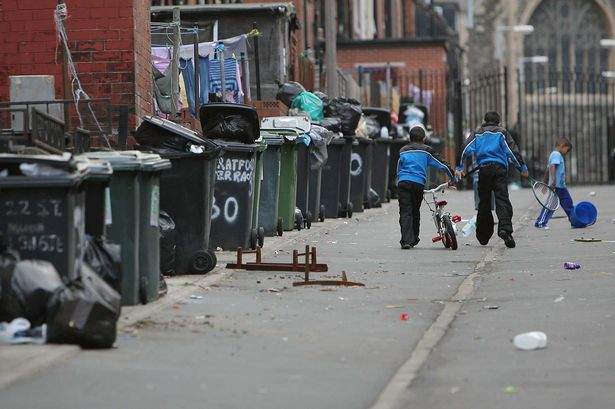 Overflowing rubbish bins line a residential street in the UK as two children walk past with a bicycle.
