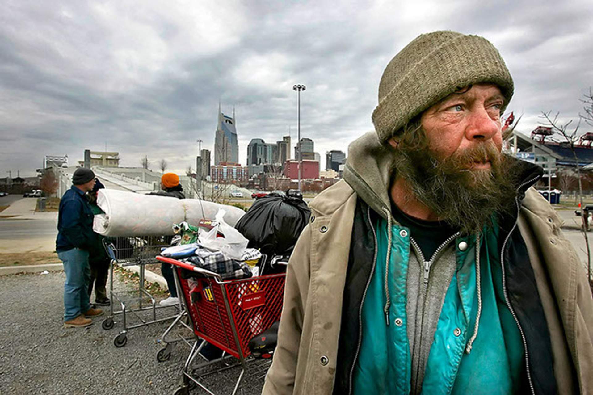 A homeless man with a beard stands beside overloaded shopping carts, with the Nashville skyline in the background.