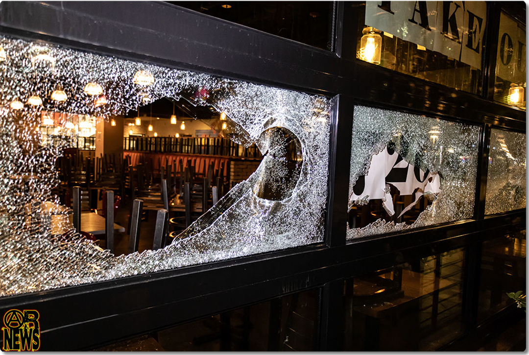 A restaurant's takeout window area with a large jagged hole smashed through the glass, tables and string lights visible inside. [Radical Edward - Untelevised Media]