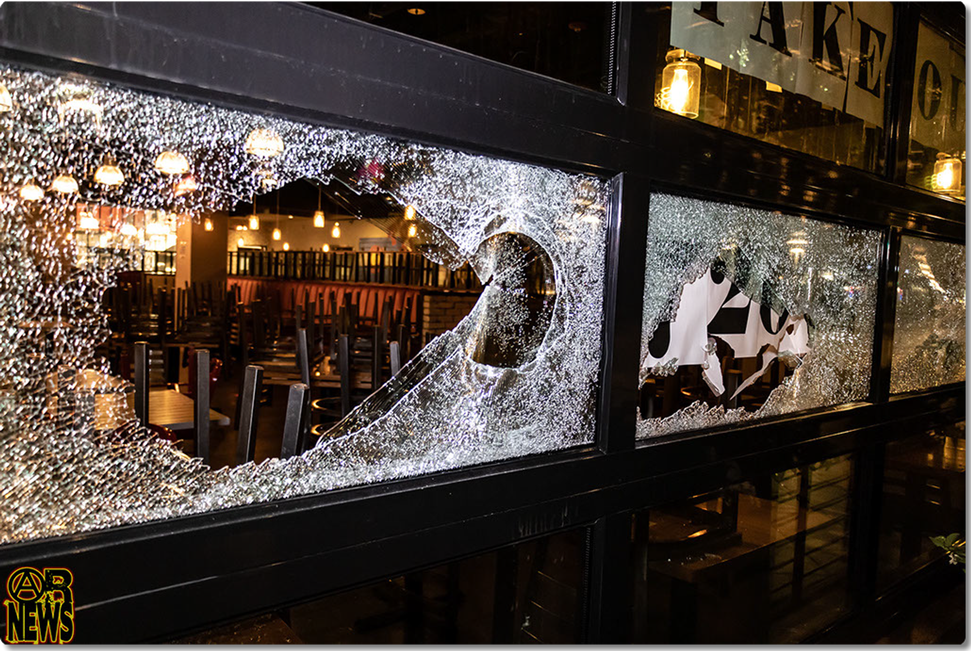 A restaurant's takeout window area with a large jagged hole smashed through the glass, tables and string lights visible inside. [Radical Edward - Untelevised Media]
