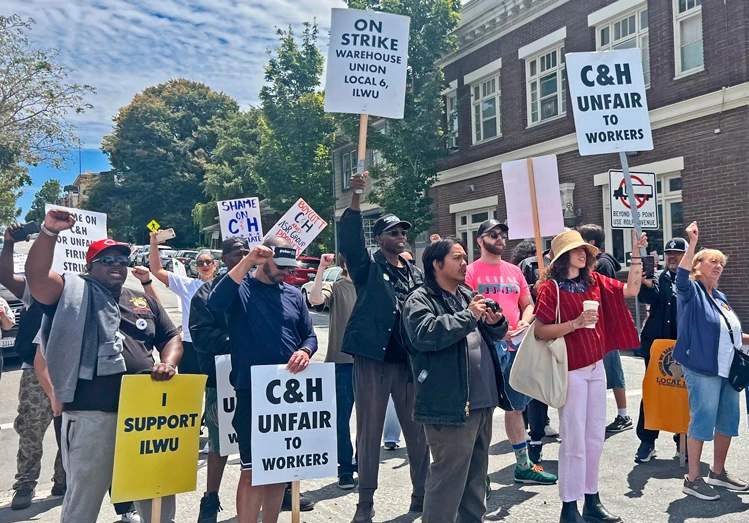 Striking workers hold ILWU union flags and signs on a street near a refinery entrance during the Sugartown Festival in Crockett California