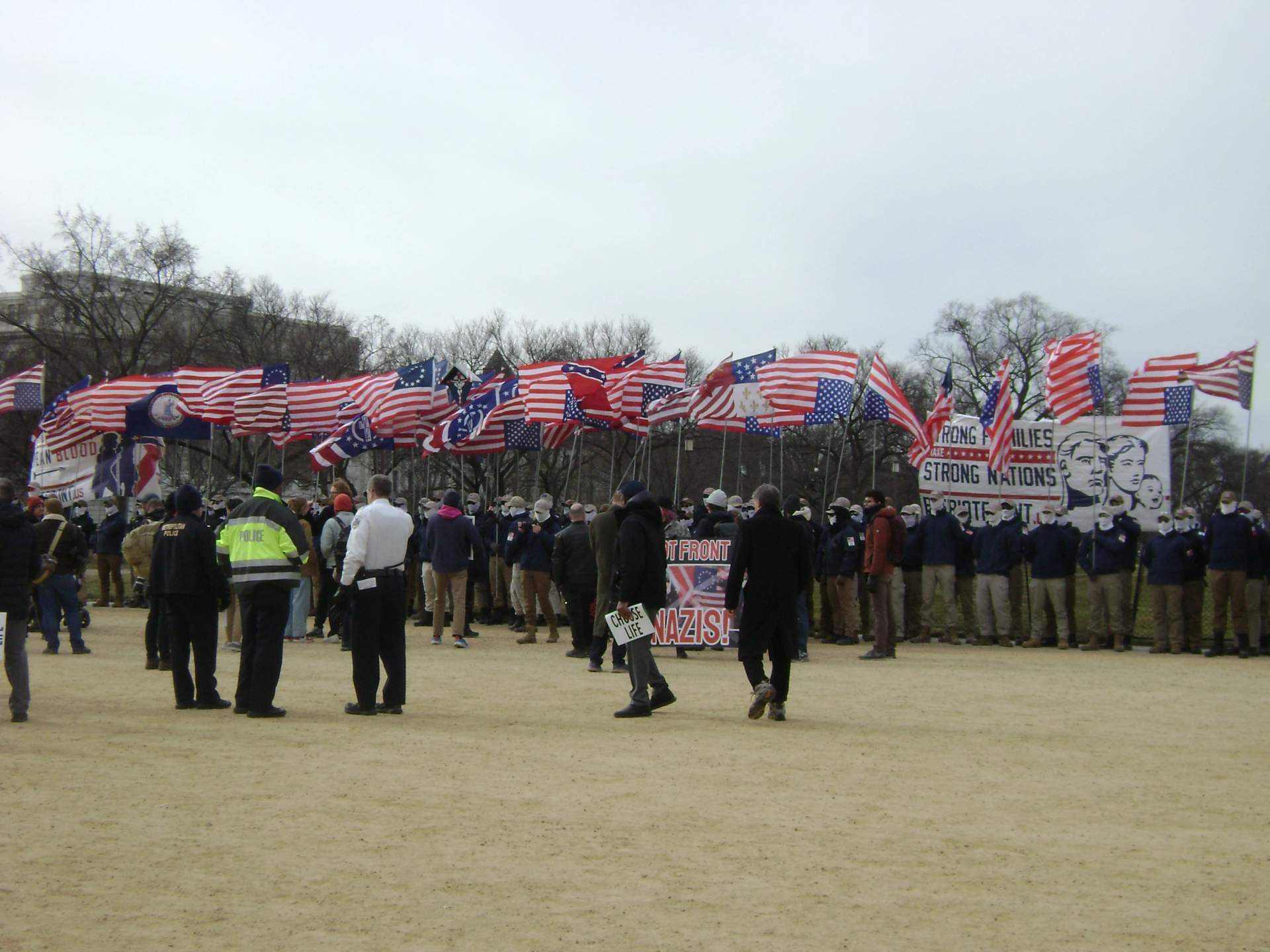 Rows of masked men in matching khaki outfits carrying flags and shields marching down a city street in formation