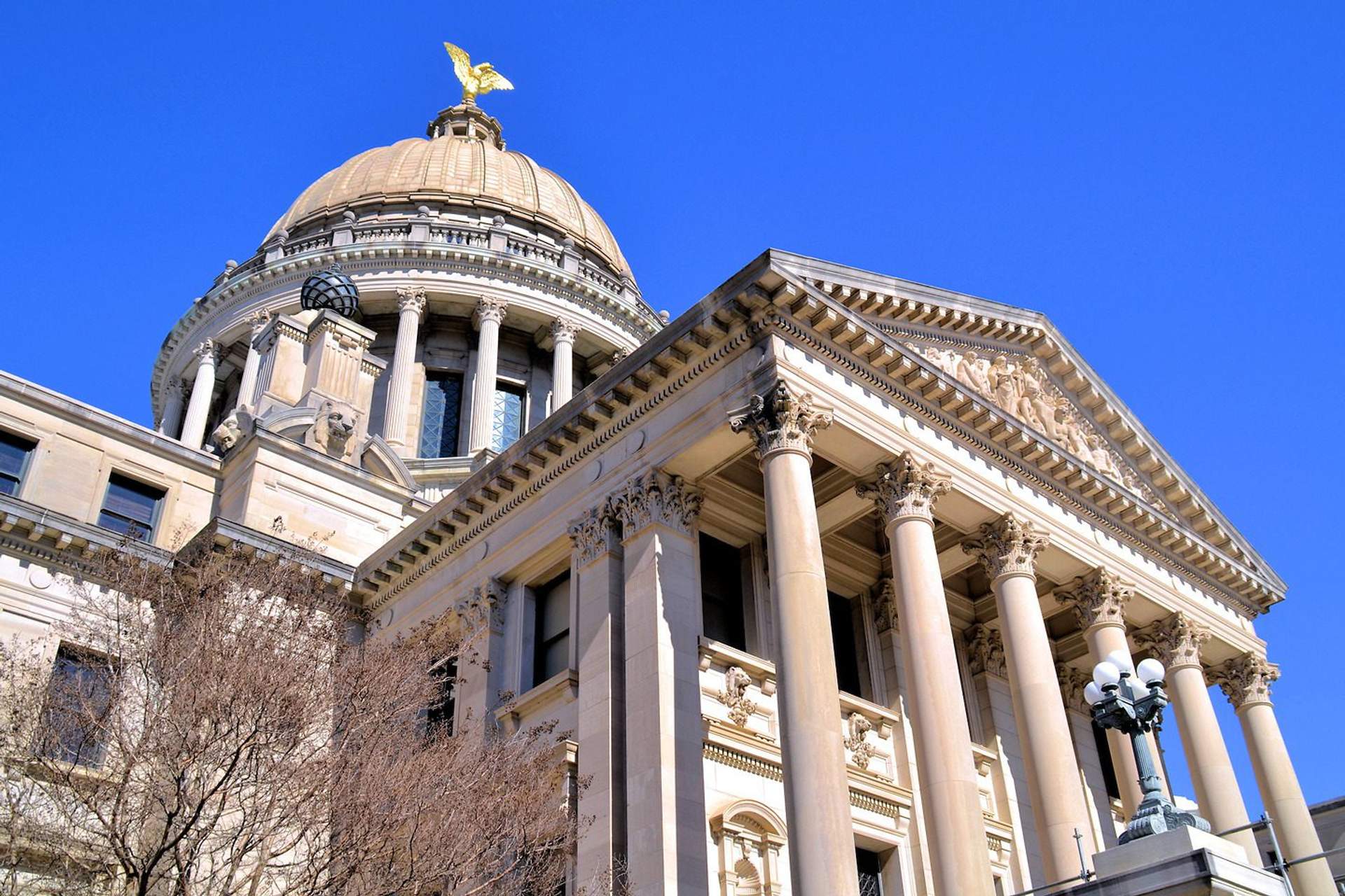 The Mississippi State Capitol building, a grand neoclassical structure with a large dome and columns, stands under a blue sky with white clouds. The building is made of light-colored stone and features American and Mississippi flags flying in front. It sits on a manicured lawn with trees framing the composition.