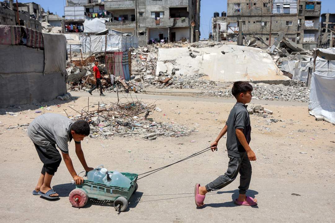 Palestinian children pushing a water cart through destroyed displacement camp in Gaza City past tents and rubble