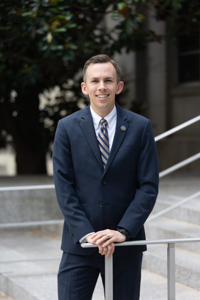 Shad White, a white man in his late 30s with dark hair and glasses, wears a dark suit and stands against a plain background with his arms crossed. He looks directly at the camera with a serious expression. This is an official portrait from his campaign website.