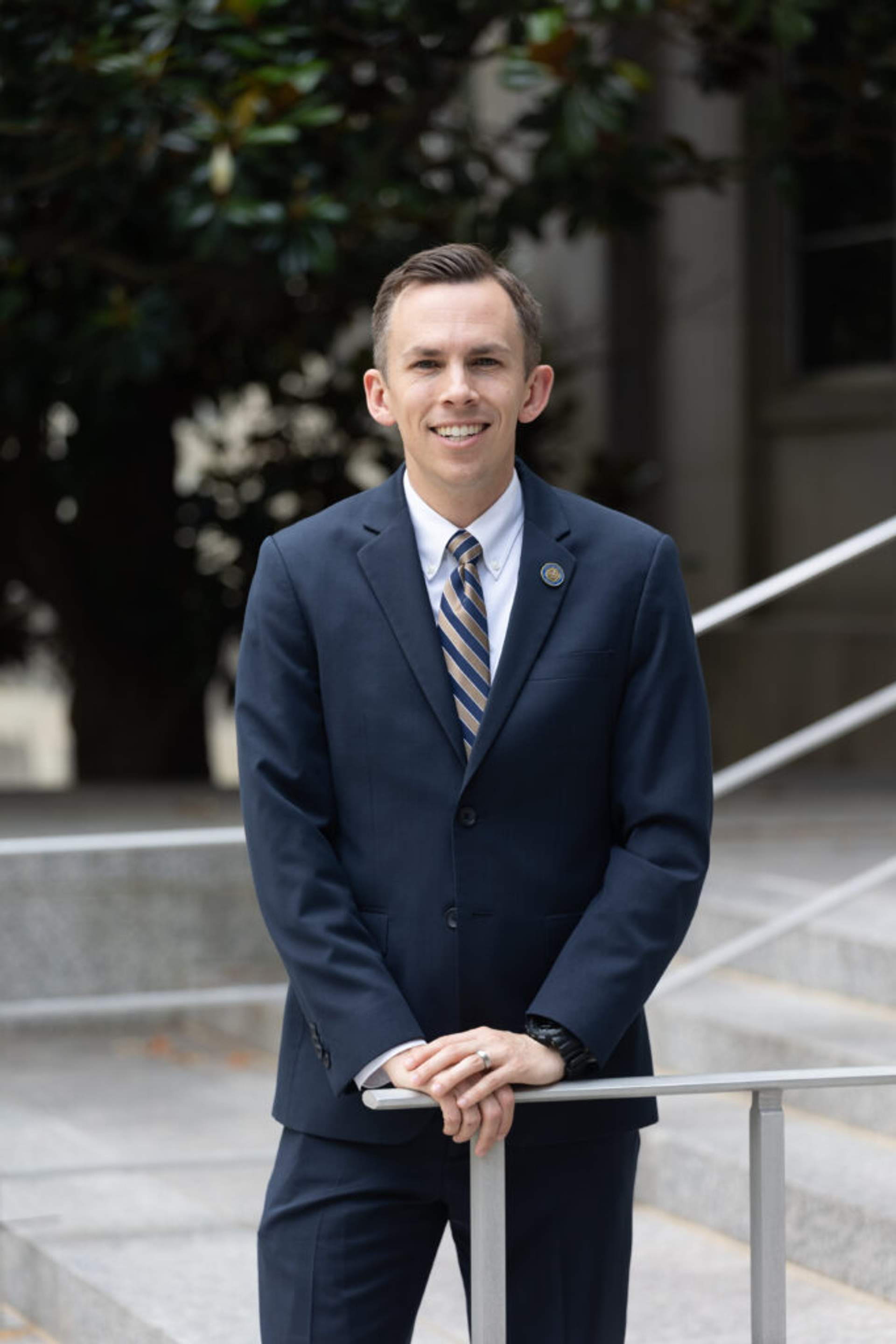 Shad White, a white man in his late 30s with dark hair and glasses, wears a dark suit and stands against a plain background with his arms crossed. He looks directly at the camera with a serious expression. This is an official portrait from his campaign website.