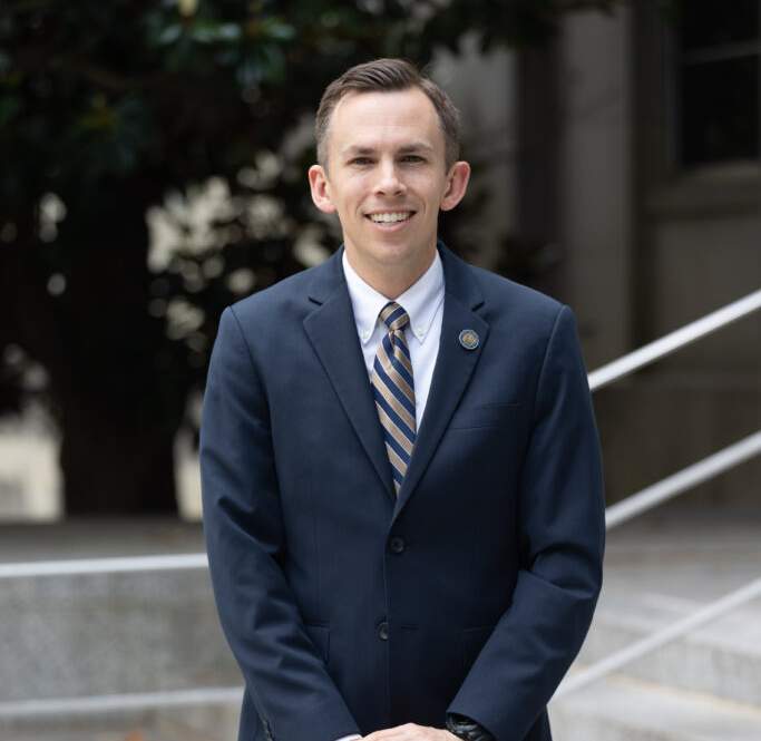 Shad White, a white man in his late 30s with dark hair and glasses, wears a dark suit and stands against a plain background with his arms crossed. He looks directly at the camera with a serious expression. This is an official portrait from his campaign website.