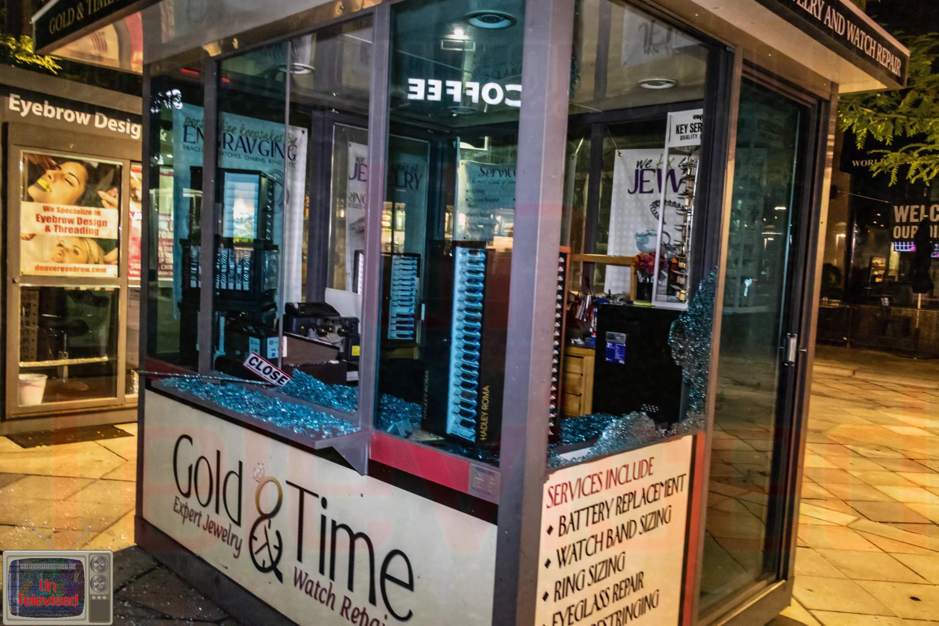 The "Gold & Time" luxury jewelry and watch repair kiosk on the mall with its display windows smashed, glass shards scattered across the counter.  [Radical Edward - Untelevised Media]
