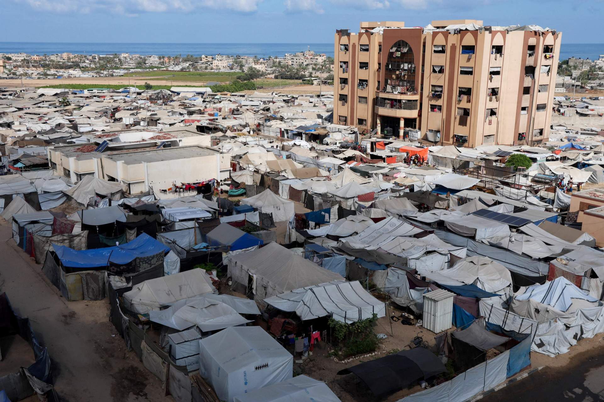 Displaced Palestinians in tent camp in Gaza with destroyed buildings visible in background