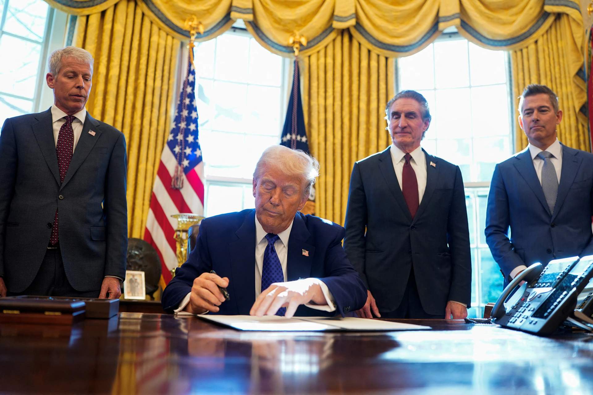 Donald Trump sitting at the Resolute Desk in the Oval Office signing a document with executive order framework visible