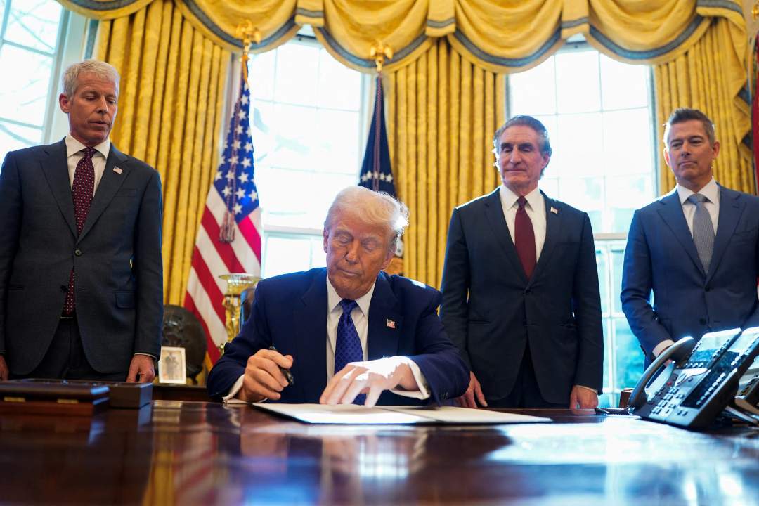 Donald Trump sitting at the Resolute Desk in the Oval Office signing a document with executive order framework visible