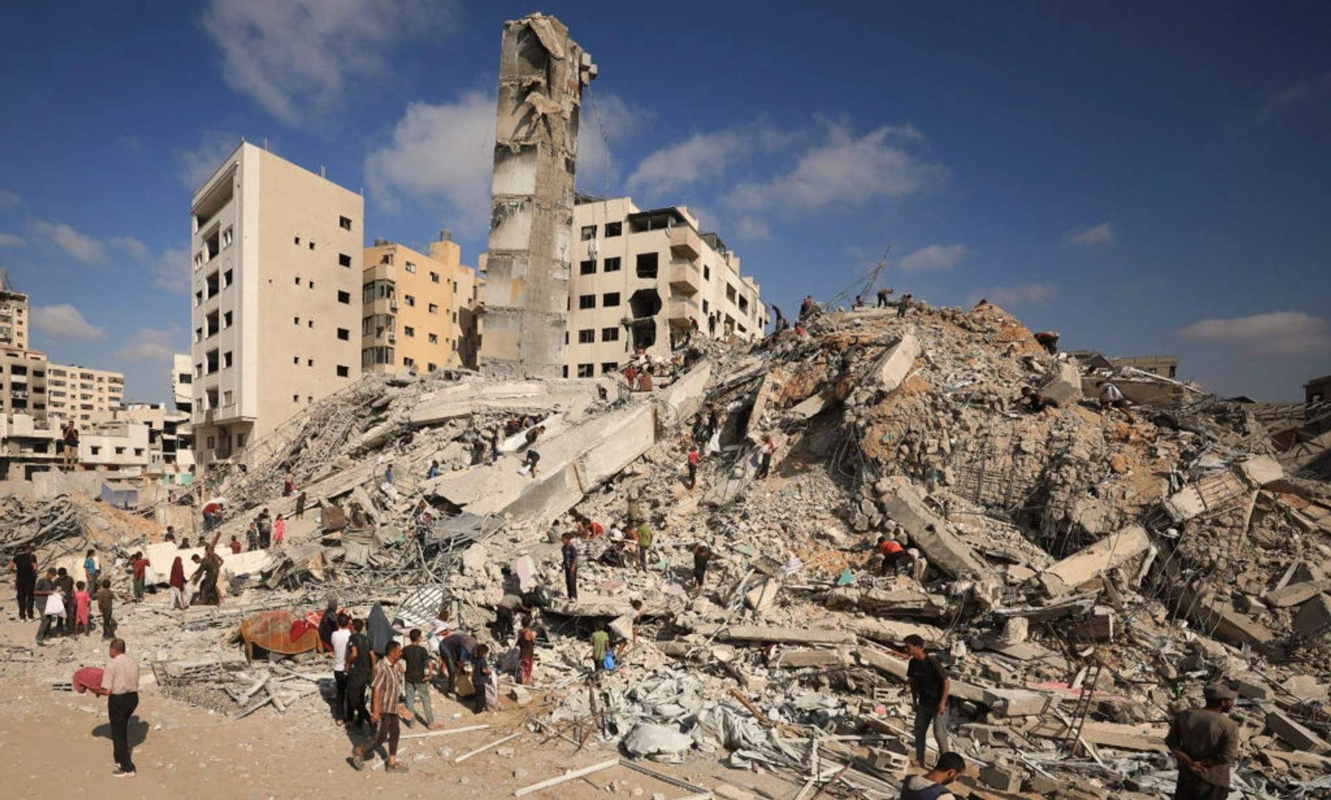 Palestinian men standing amid concrete rubble and twisted rebar from a destroyed multi-story residential building after an Israeli airstrike in Gaza City