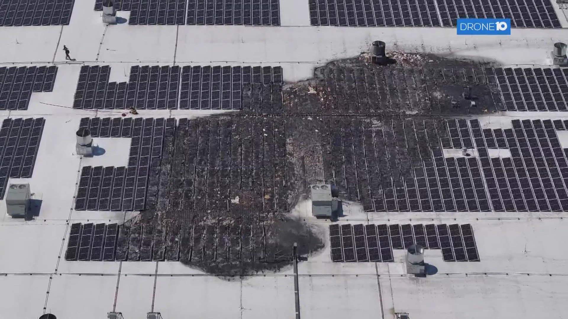 Aerial or elevated view of a large Amazon warehouse building with thick black smoke rising from the roof where solar panels are visible, with fire trucks and emergency vehicles positioned below on a clear day.