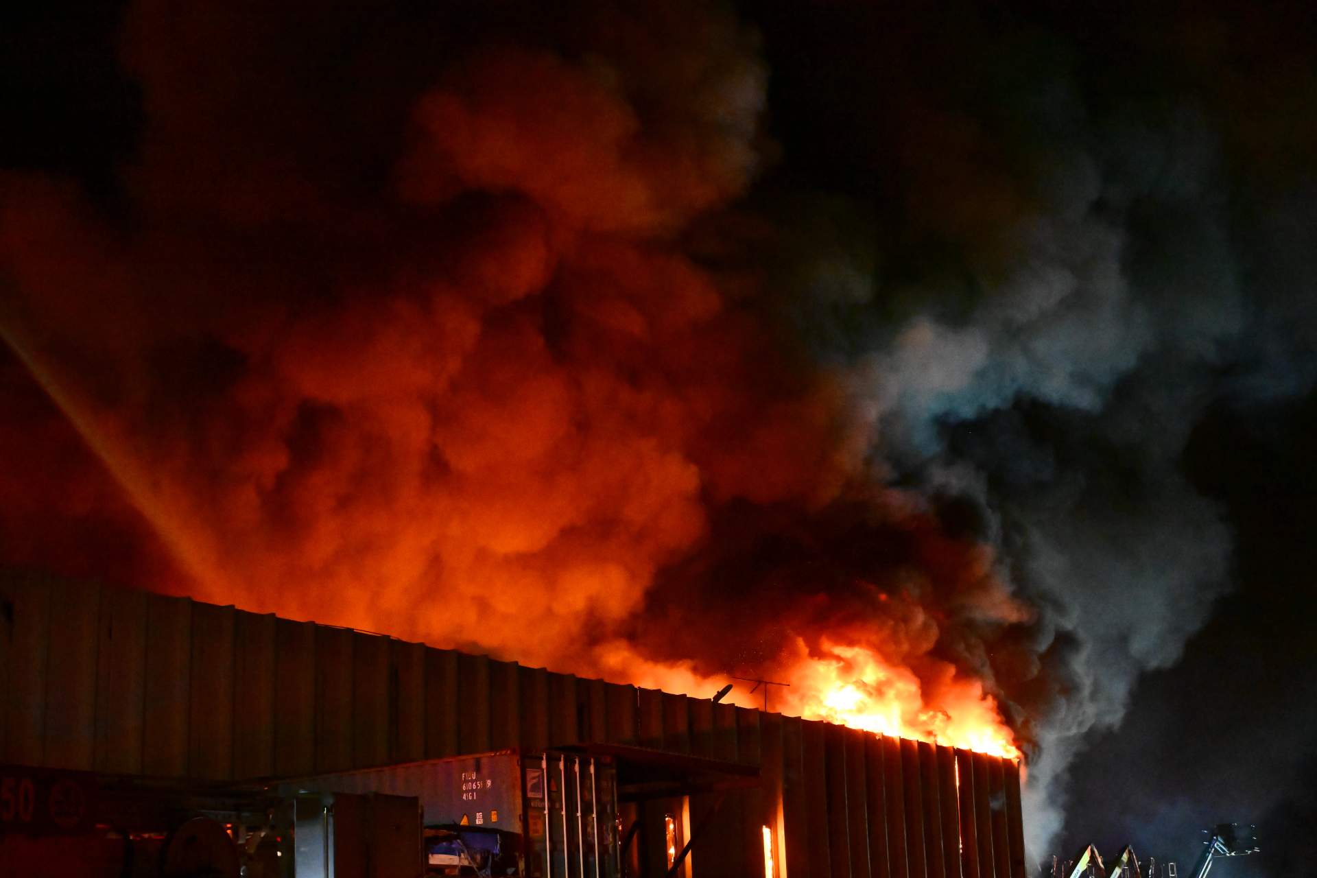 Nighttime view of a large industrial building engulfed in flames with multiple fire department tower ladders positioned around it, their water streams illuminated by emergency lights.