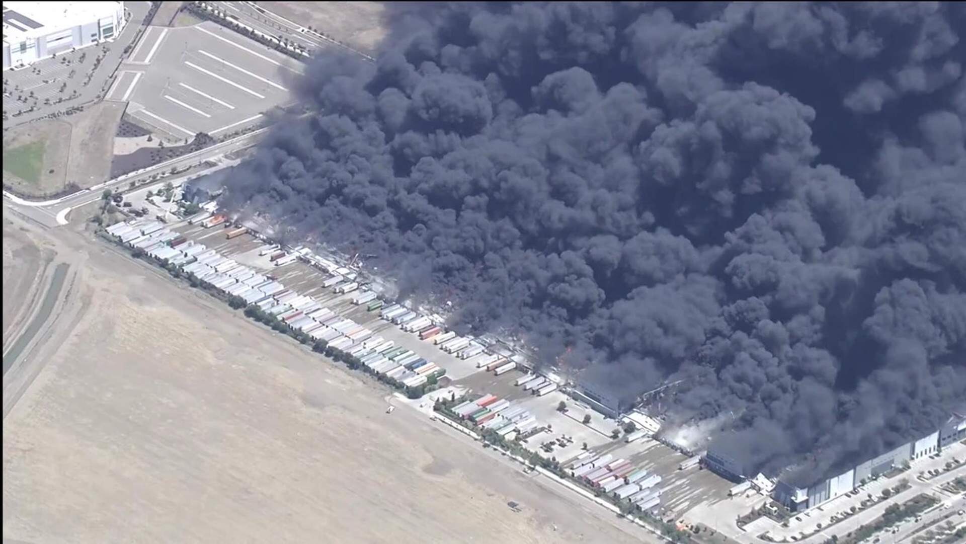 Helicopter aerial view showing a massive warehouse fire at the Medline Industries distribution center with thick black smoke billowing across the sky in Tracy, California.