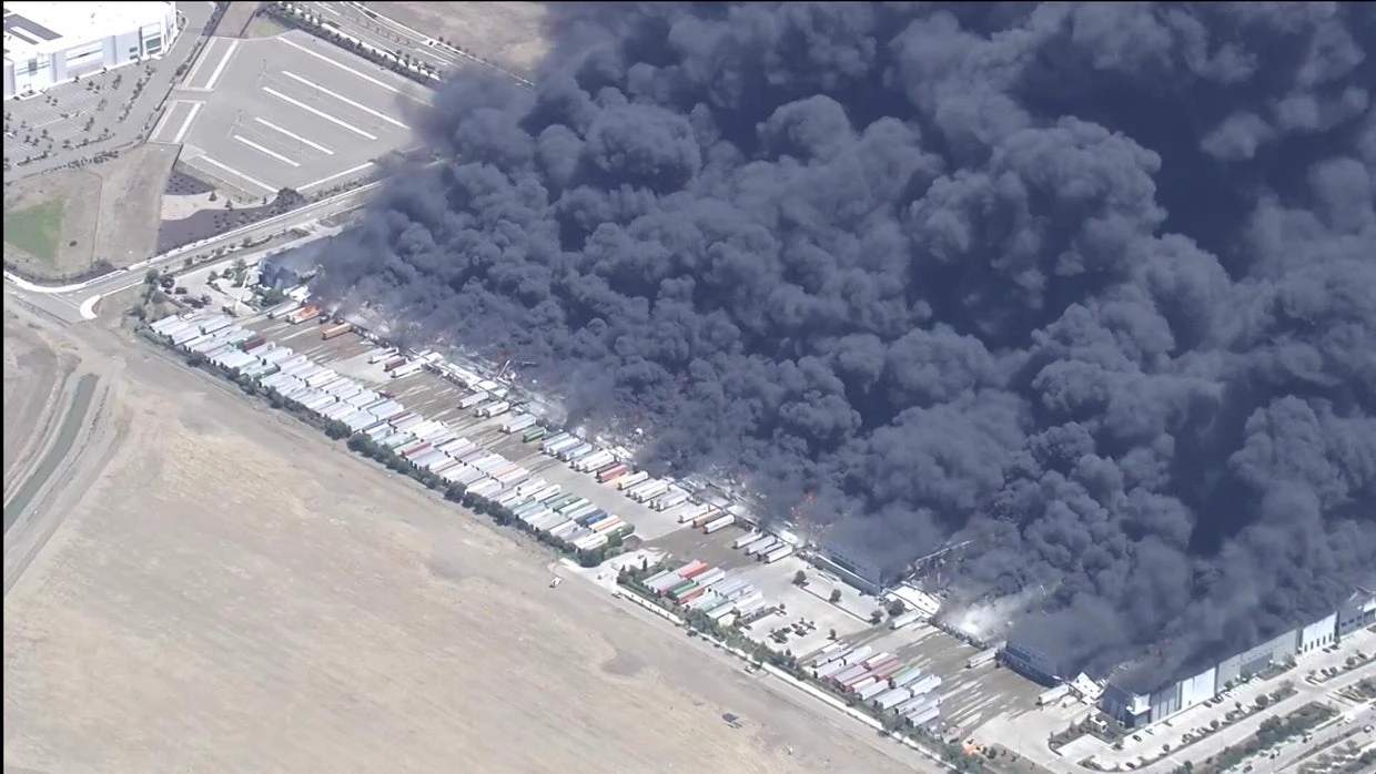 Helicopter aerial view showing a massive warehouse fire at the Medline Industries distribution center with thick black smoke billowing across the sky in Tracy, California.