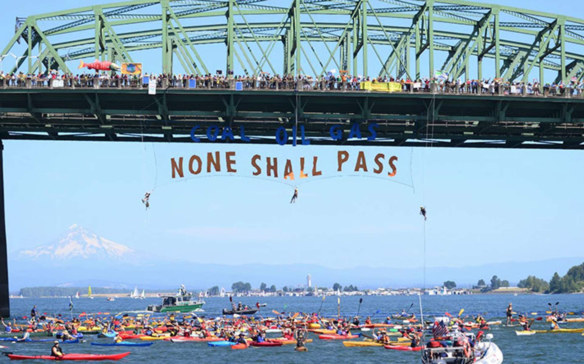 Activists suspended beneath the St. Johns Bridge display a banner reading 'Coal Oil Gas — None Shall Pass' over a flotilla of kayaks, with Mount Hood visible in the distance.