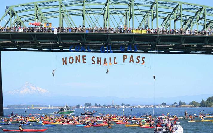 Activists suspended beneath the St. Johns Bridge display a banner reading 'Coal Oil Gas — None Shall Pass' over a flotilla of kayaks, with Mount Hood visible in the distance.