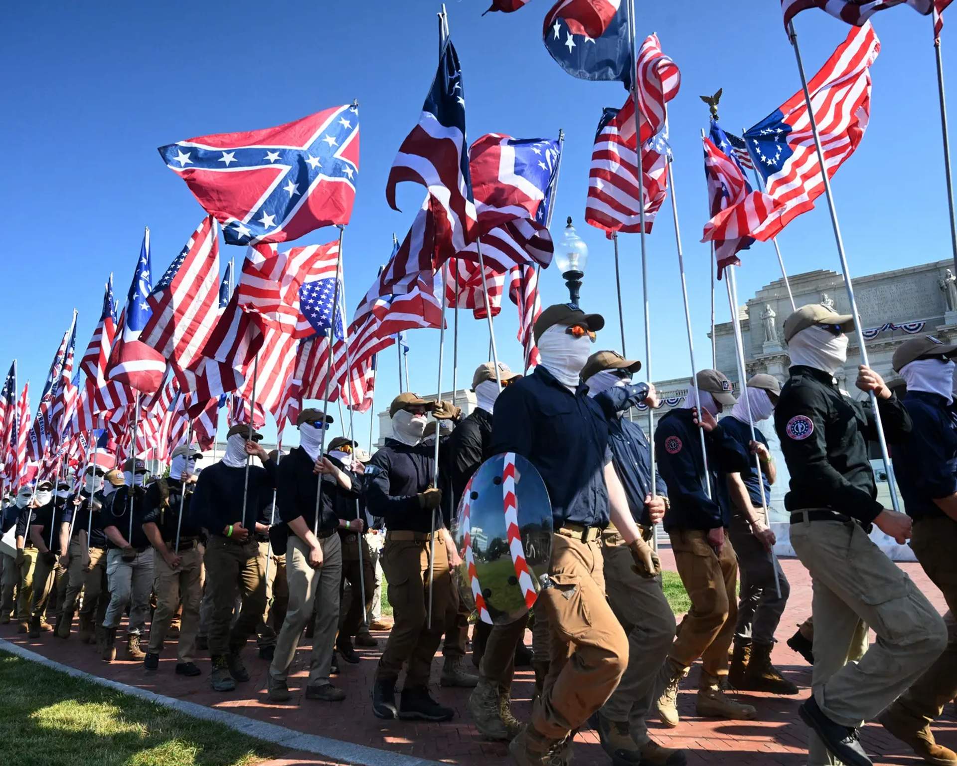 Masked men in navy shirts march in formation down a sidewalk carrying American and Confederate flags with the US Capitol visible in the background