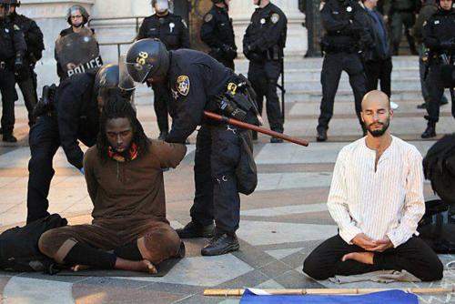 Riot police restrain and search a seated protester while another protester nearby sits calmly in meditation.