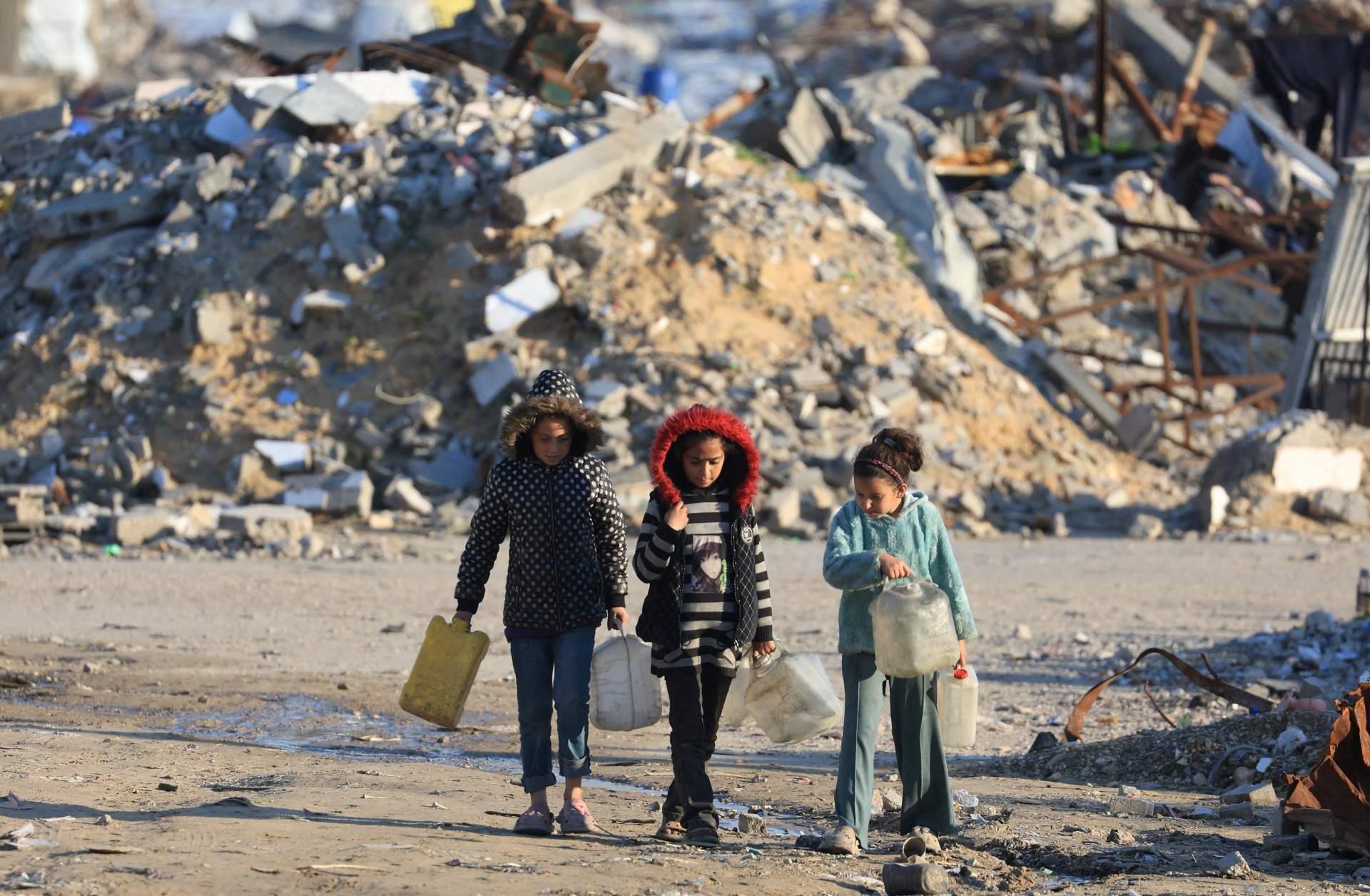 Two Palestinian girls walking past towering piles of concrete rubble from destroyed multi-story buildings in Gaza during the ceasefire period