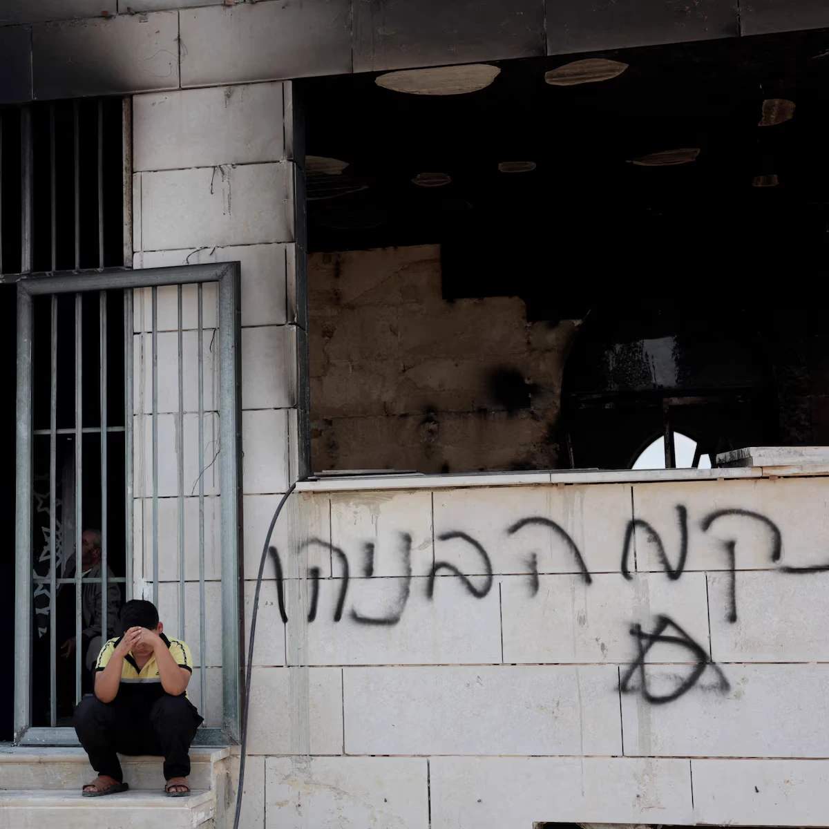 Palestinian sitting on the steps outside a damaged mosque with visible fire and graffiti damage
