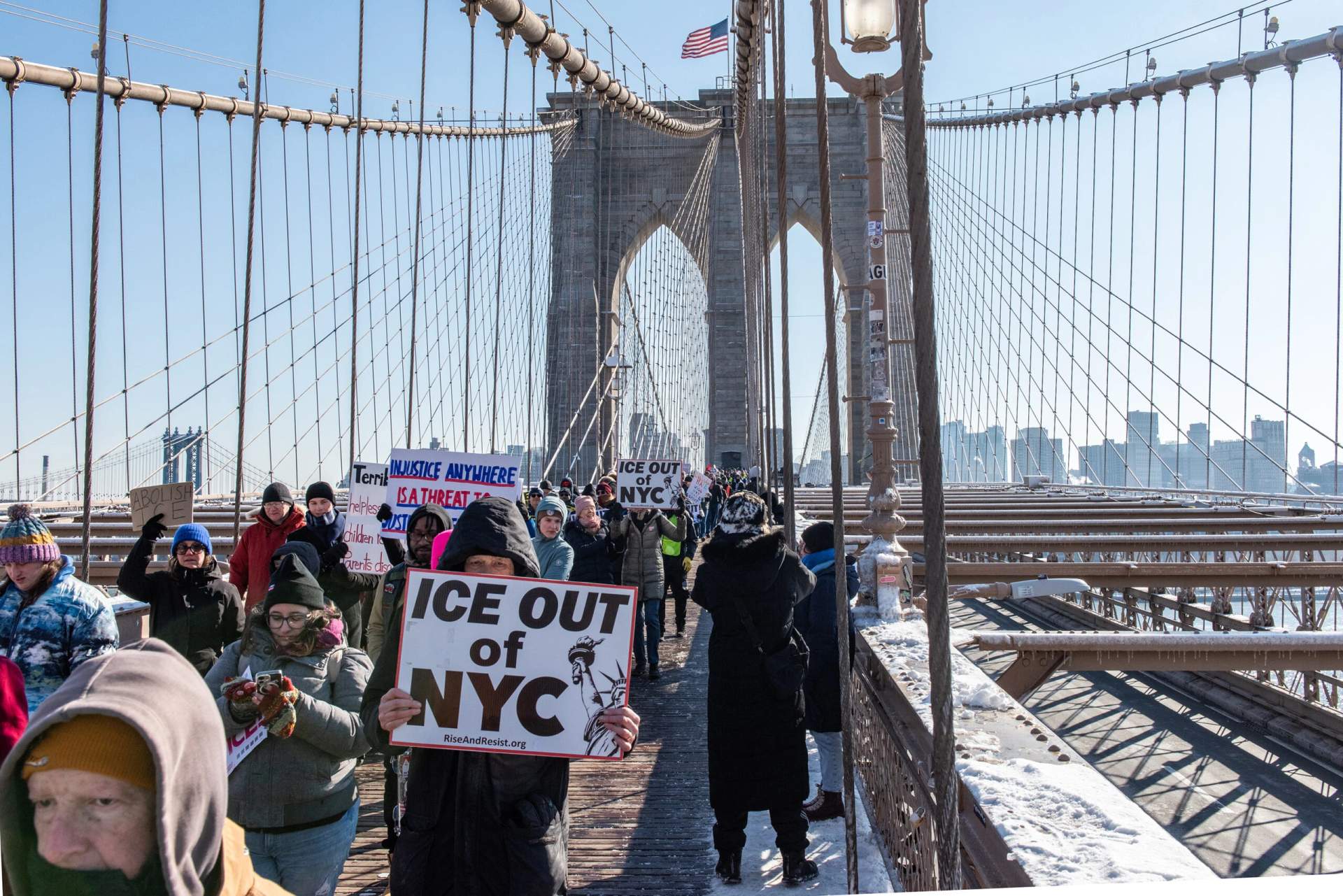 Large crowd of protesters marching across the Brooklyn Bridge carrying signs and banners during an anti-ICE demonstration on January 19, 2026