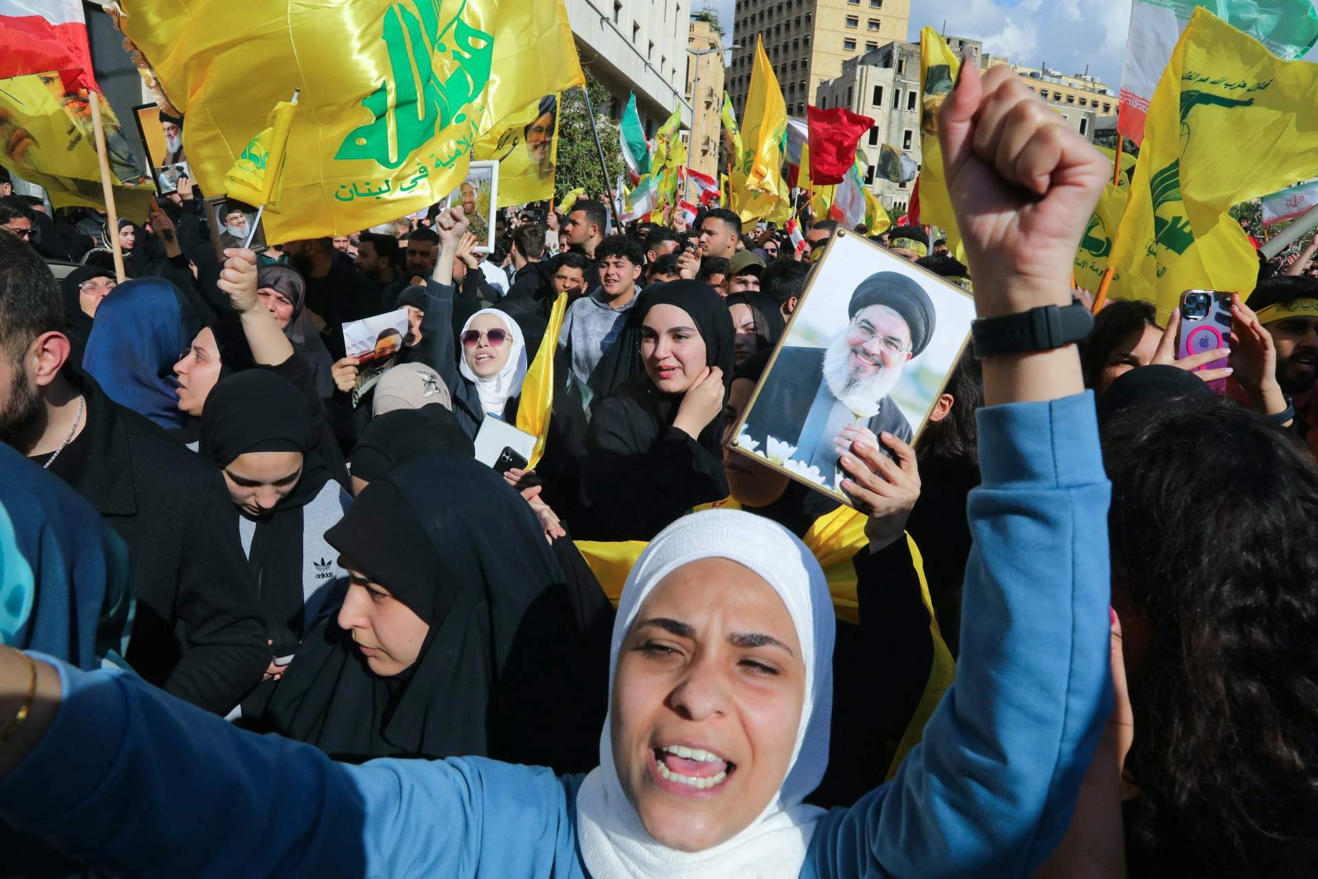 Crowd of Hezbollah supporters at a protest in Beirut, waving yellow Hezbollah flags and holding up printed images, with some people raising their fists in defiance.