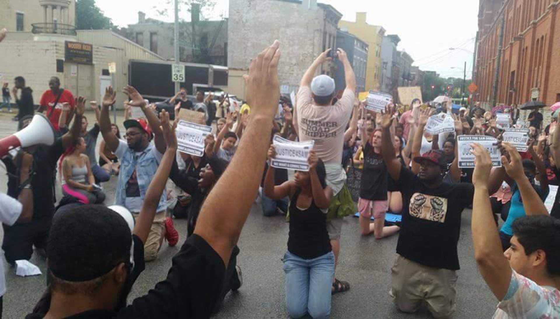 Protesters kneel in a Cincinnati street with fists raised, holding signs reading 'Justice for Sam' during a demonstration against police brutality.