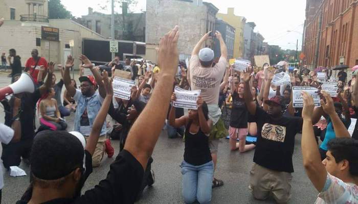 Protesters kneel in a Cincinnati street with fists raised, holding signs reading 'Justice for Sam' during a demonstration against police brutality.