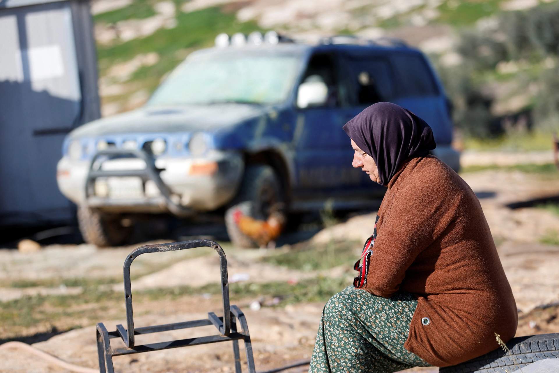 A Palestinian woman sitting on the ground next to a damaged vehicle near rural hills in the occupied West Bank