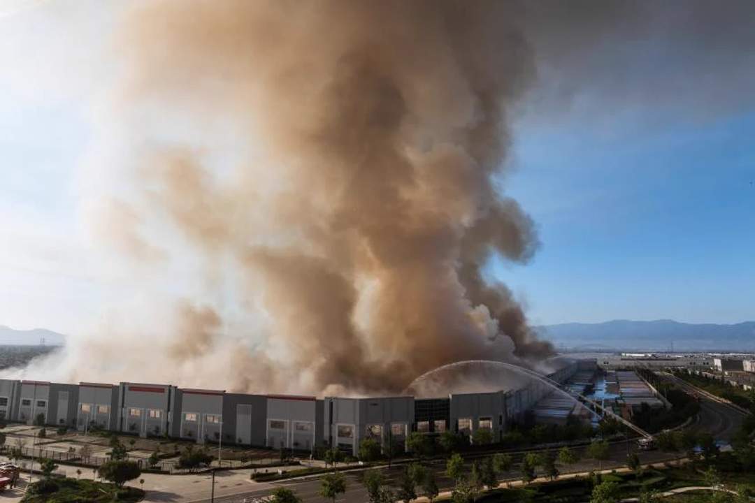 Aerial photograph of a massive industrial warehouse building consumed by bright orange flames, with thick black smoke billowing into the night sky