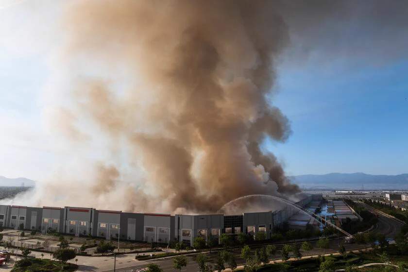 Aerial photograph of a massive industrial warehouse building consumed by bright orange flames, with thick black smoke billowing into the night sky