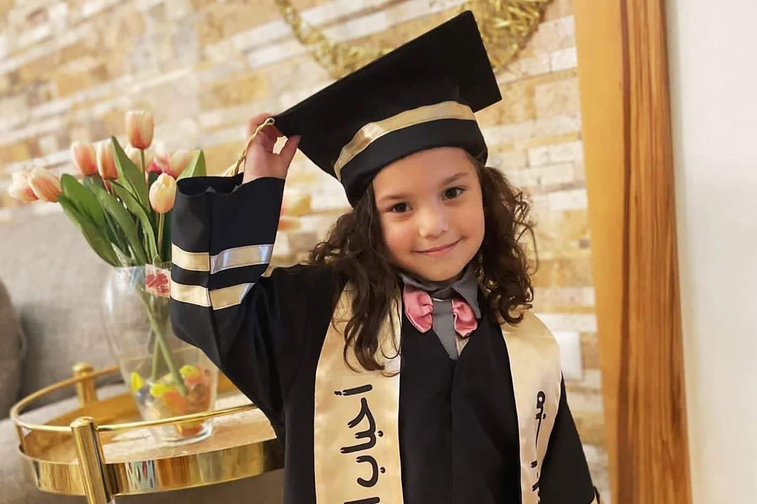 Hind Rajab in a graduation cap and gown with a gold Arabic-lettered sash, adjusting her cap tassel and smiling.