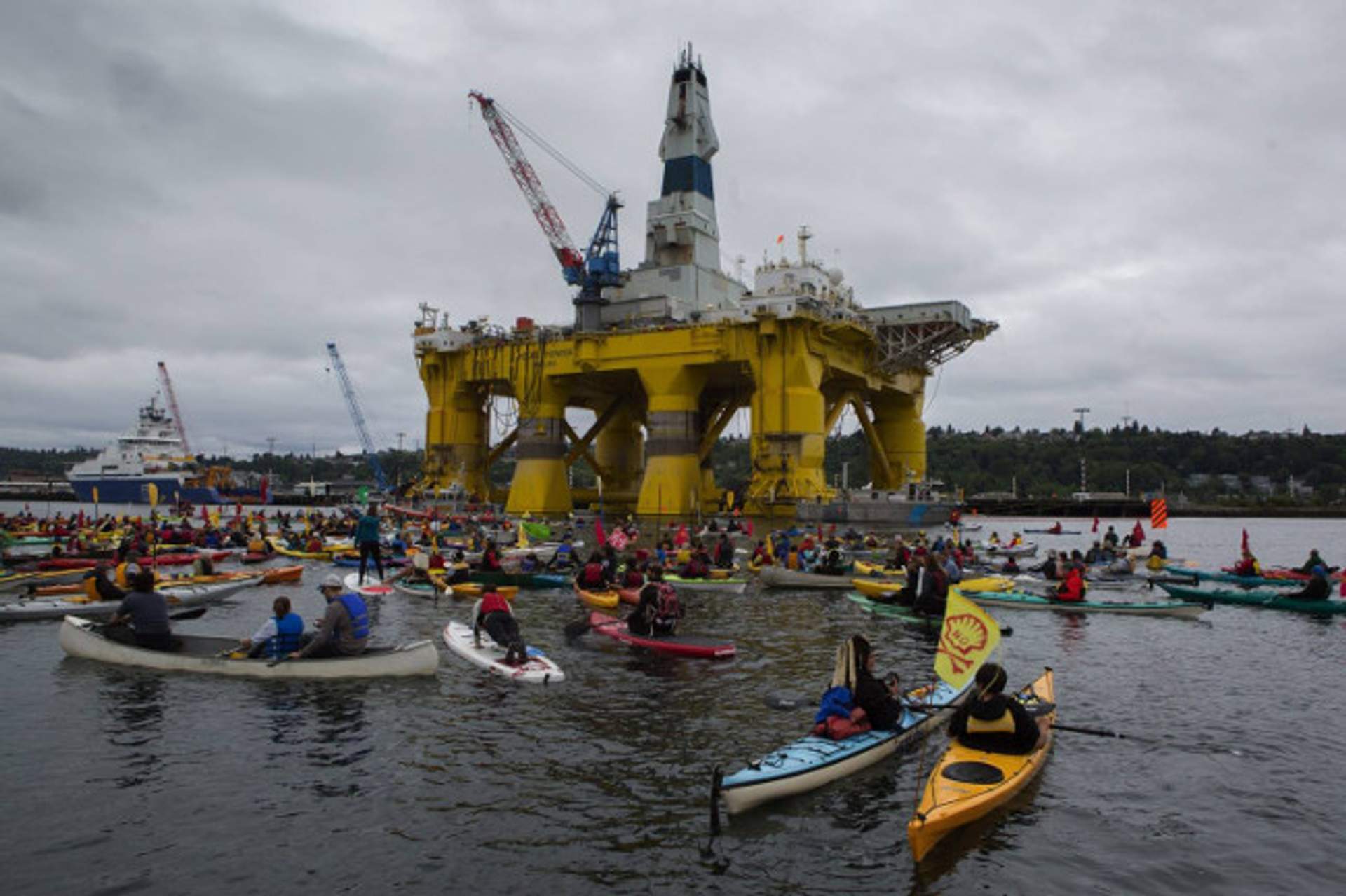 A large crowd of kayakers on the water, with the yellow Polar Pioneer rig visible in the background.