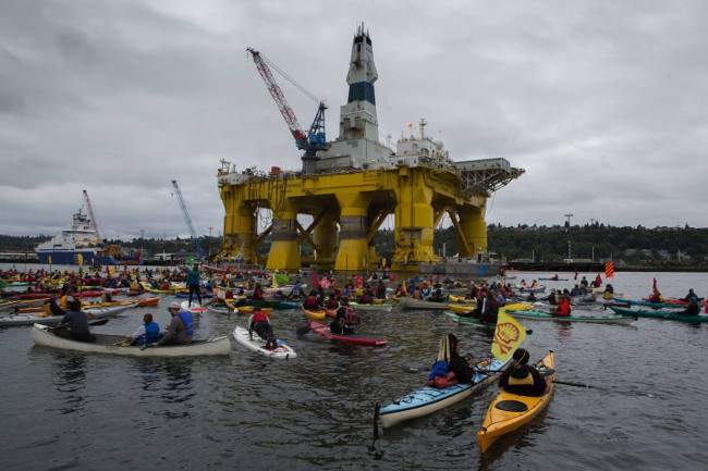 A large crowd of kayakers on the water, with the yellow Polar Pioneer rig visible in the background.