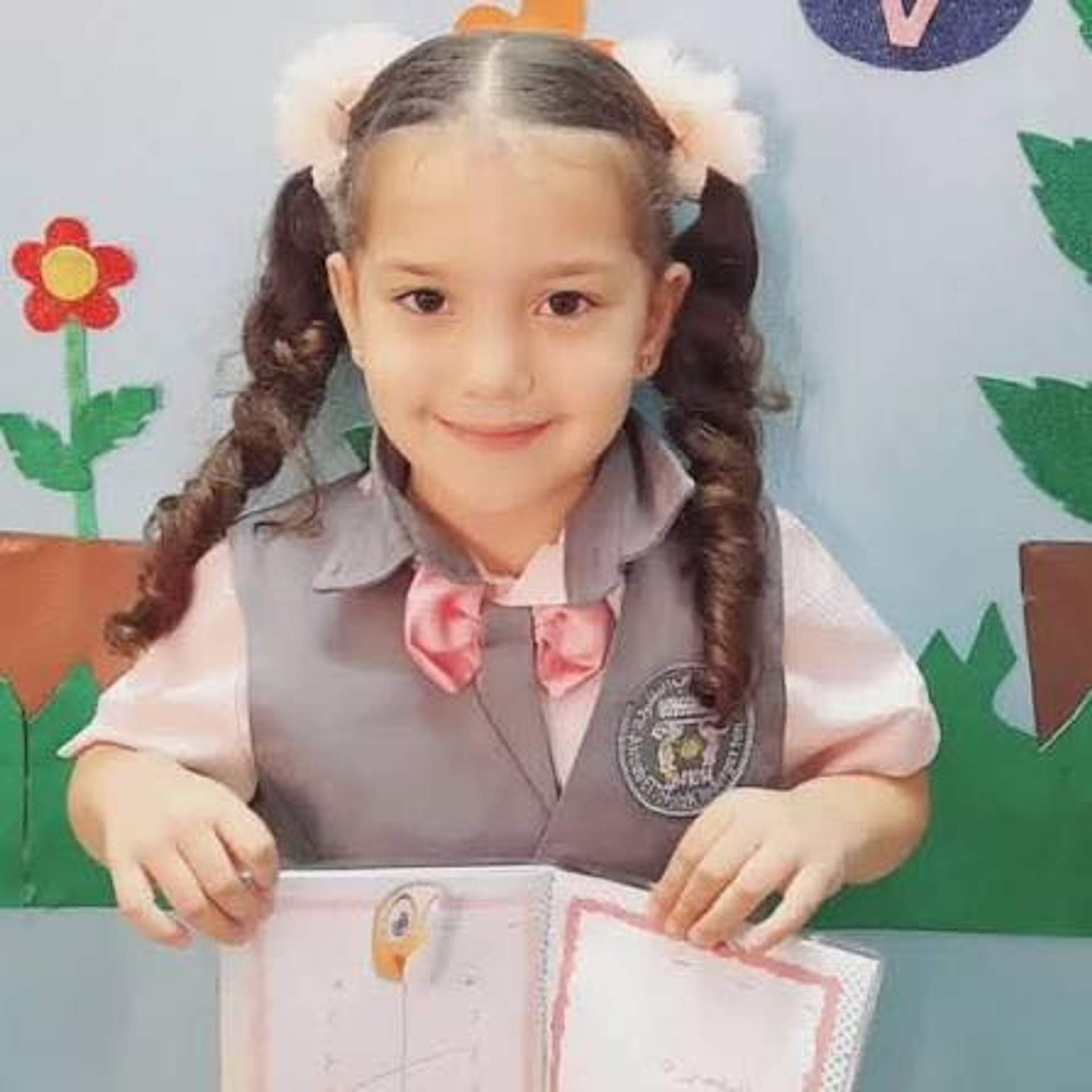 Hind Rajab in her school uniform — grey vest, pink shirt, pink bow tie — holding papers and smiling in her classroom.